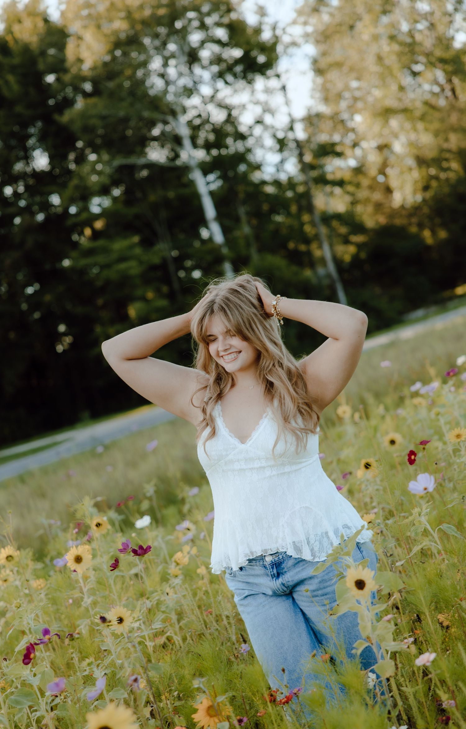 Young woman in white top and jeans smiling in a vibrant wildflower field with sunflowers and colorful blooms.