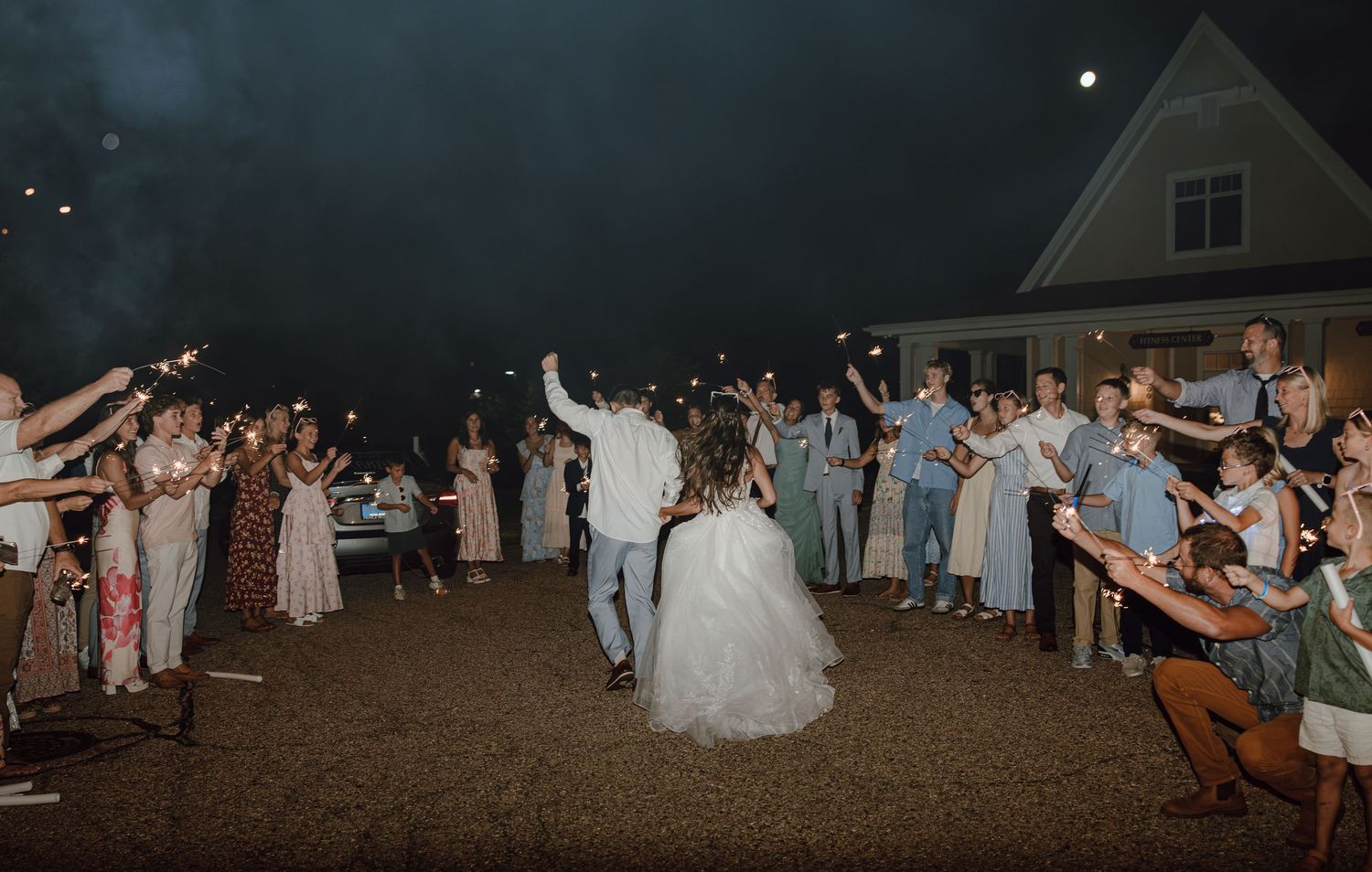 Bride and groom celebrate with sparkler send-off surrounded by cheering wedding guests at night.