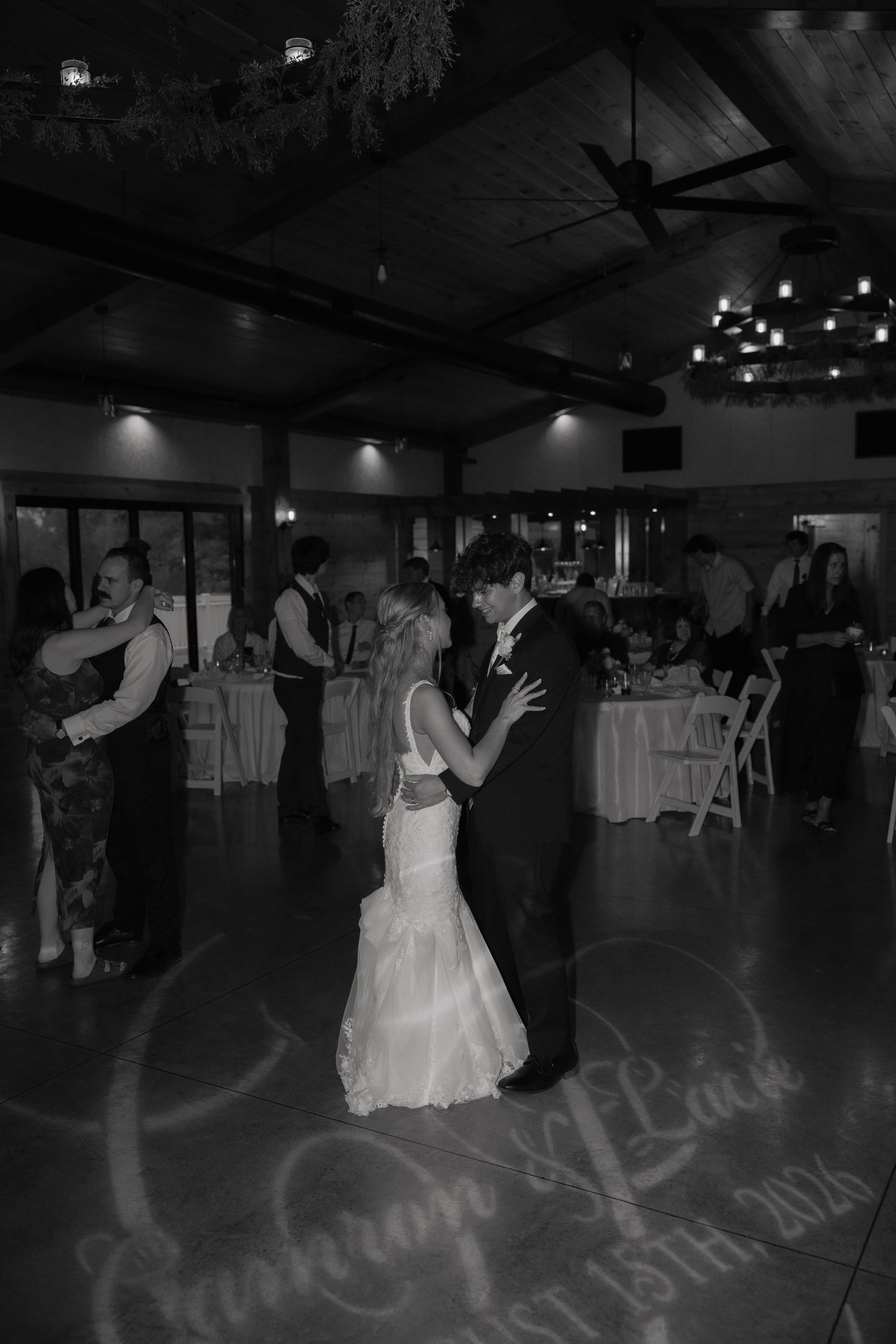 Bride and groom share their first dance at wedding reception, with names projected on dance floor in black and white photo.