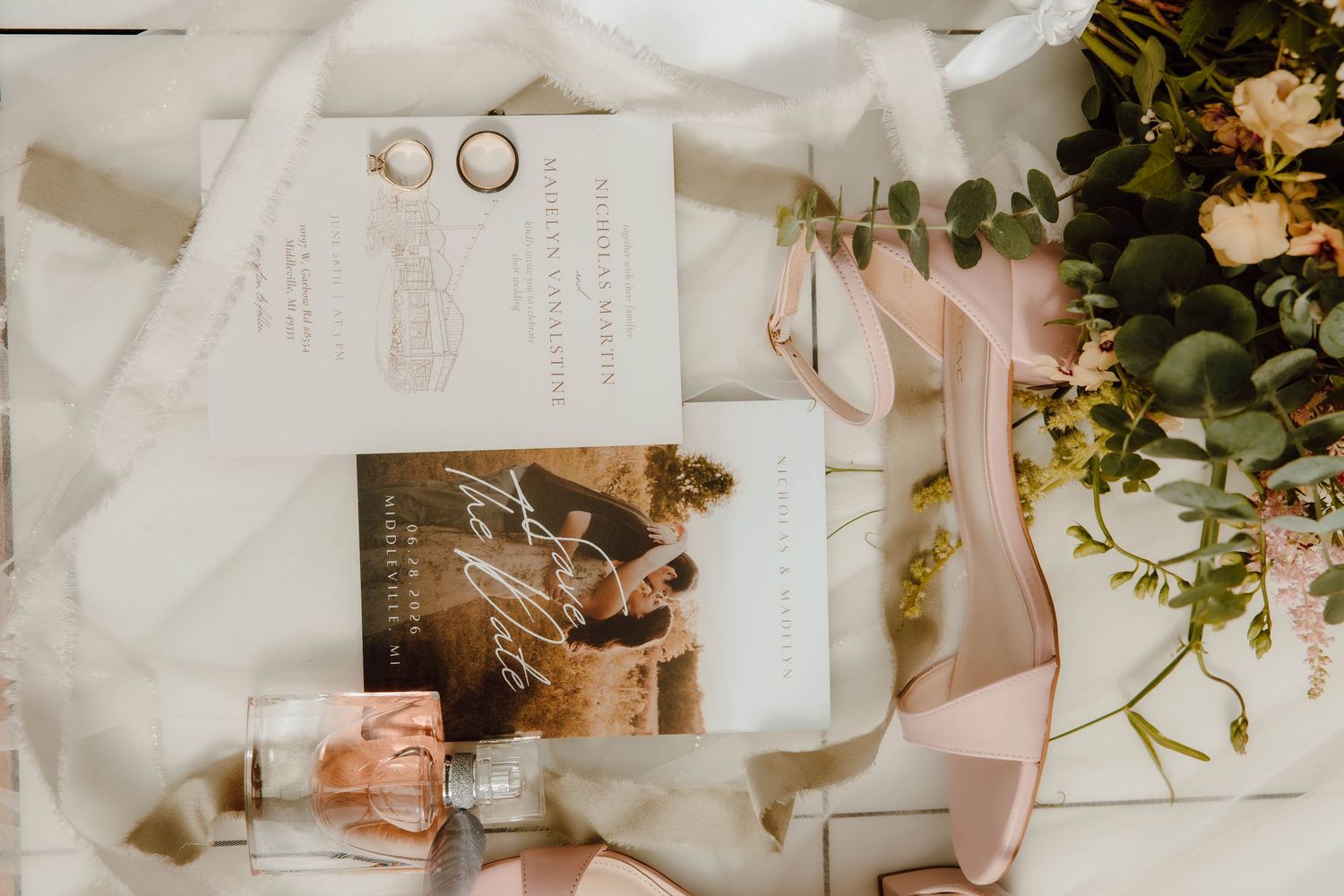 Elegant wedding flat lay with invitation suite, gold rings, blush ribbons, and floral greenery arrangement.