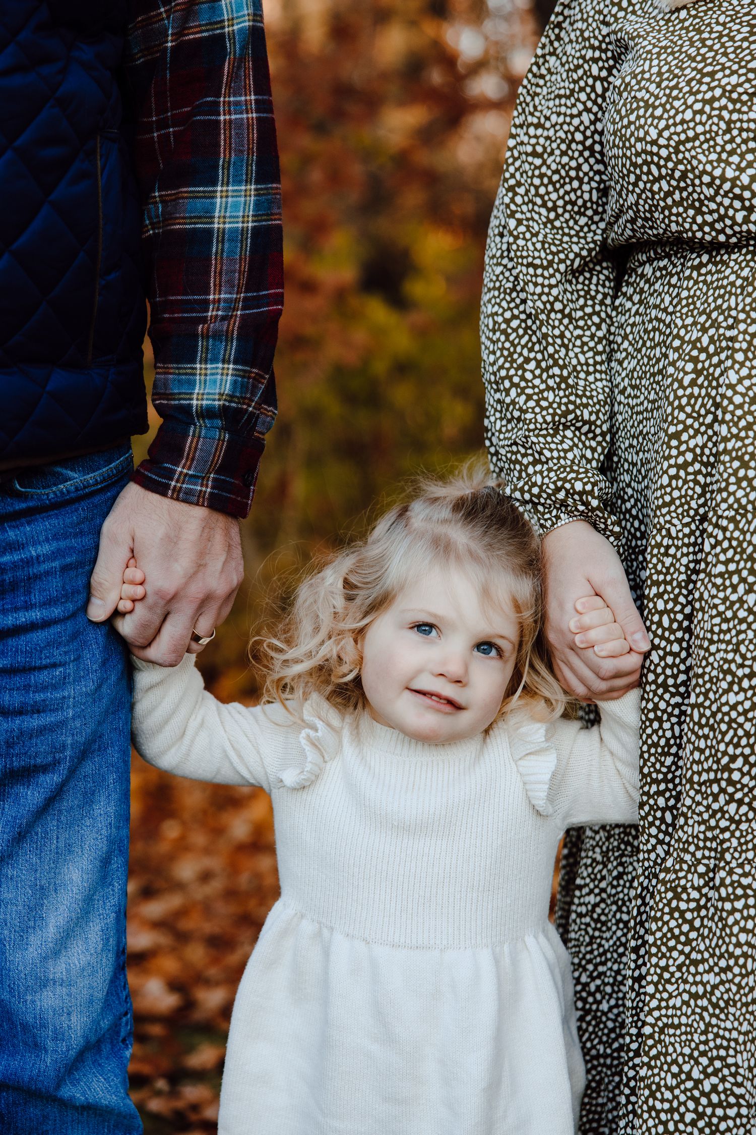 A small child in a white dress holds hands with parents during an autumn photoshoot.