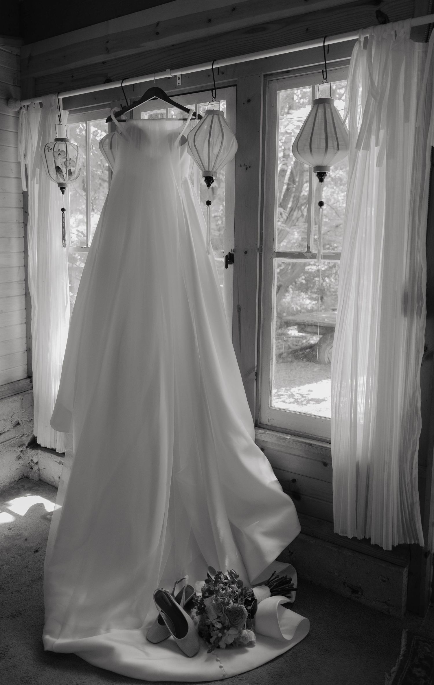 Black and white photo of an elegant wedding dress hanging by a window with bridal shoes and bouquet below.