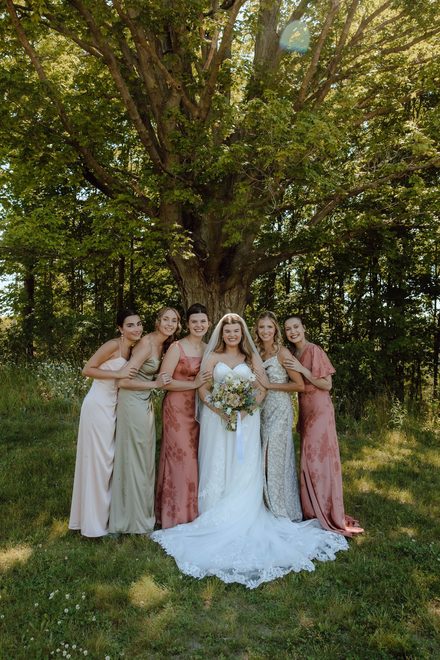 Bride in white lace gown poses with five bridesmaids in pink and sage dresses under a large tree outdoors.