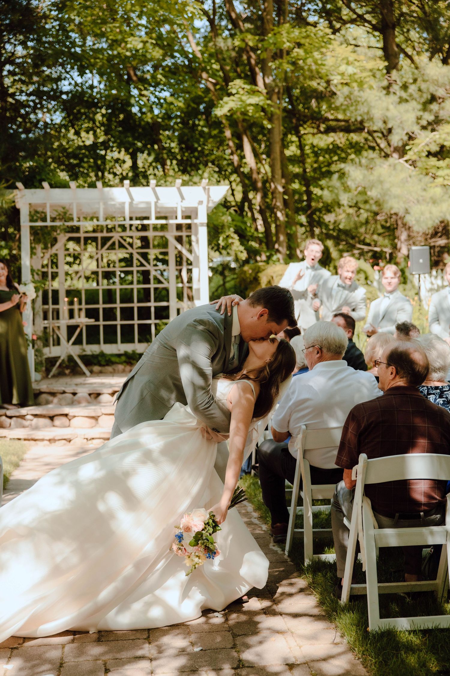 Bride and groom share a romantic dip kiss at outdoor wedding ceremony with white arbor and seated guests.