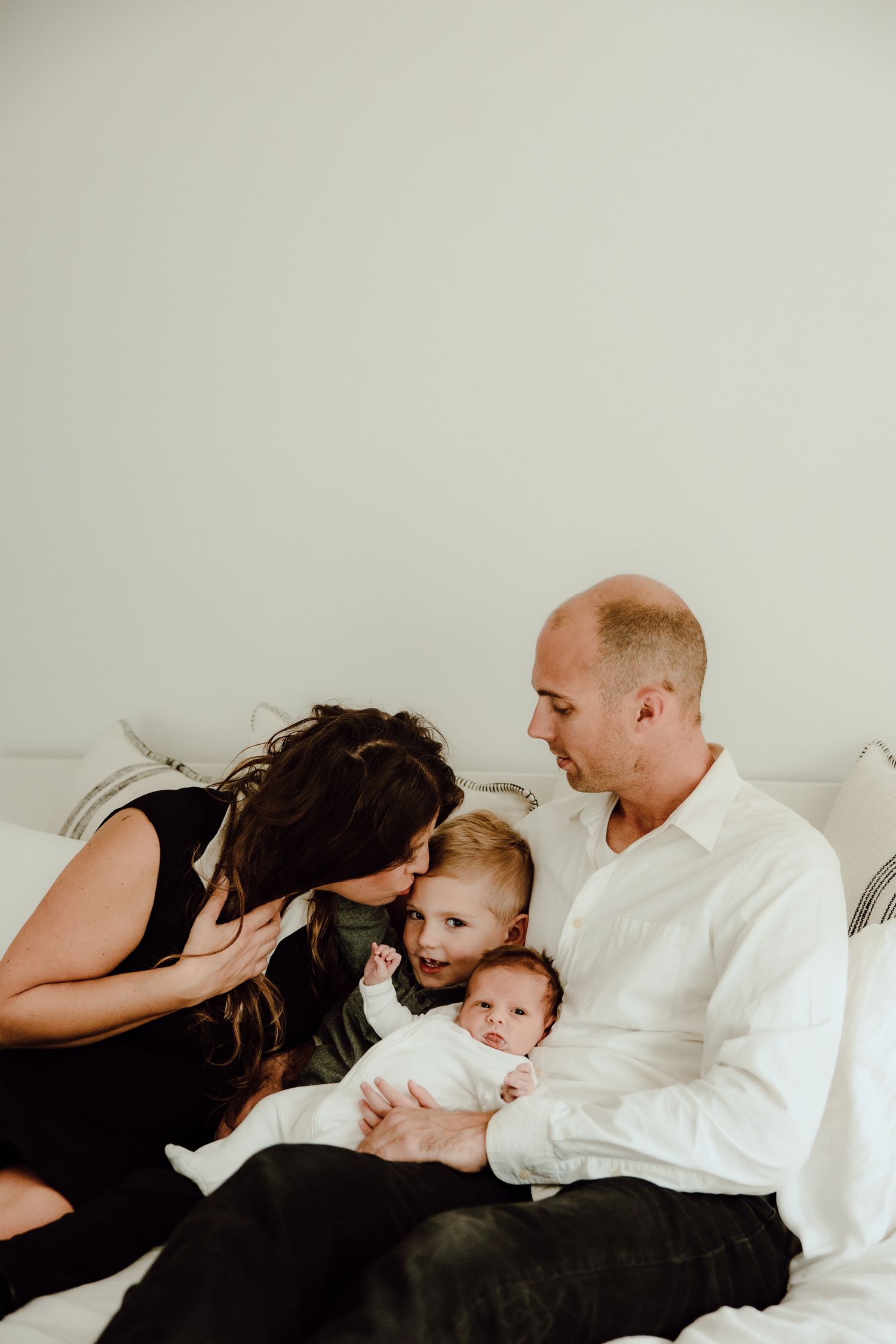 A family cuddles together on a white bed with their young children.