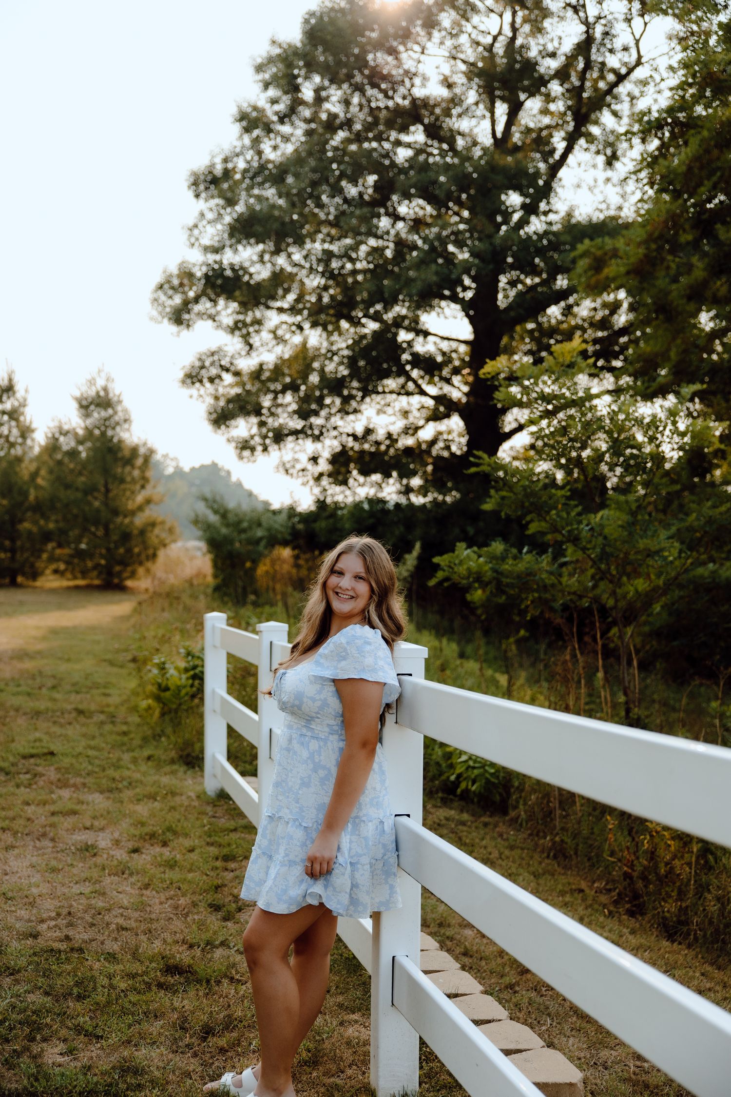 White dress contrasts against a rustic wooden fence in a pastoral countryside scene.