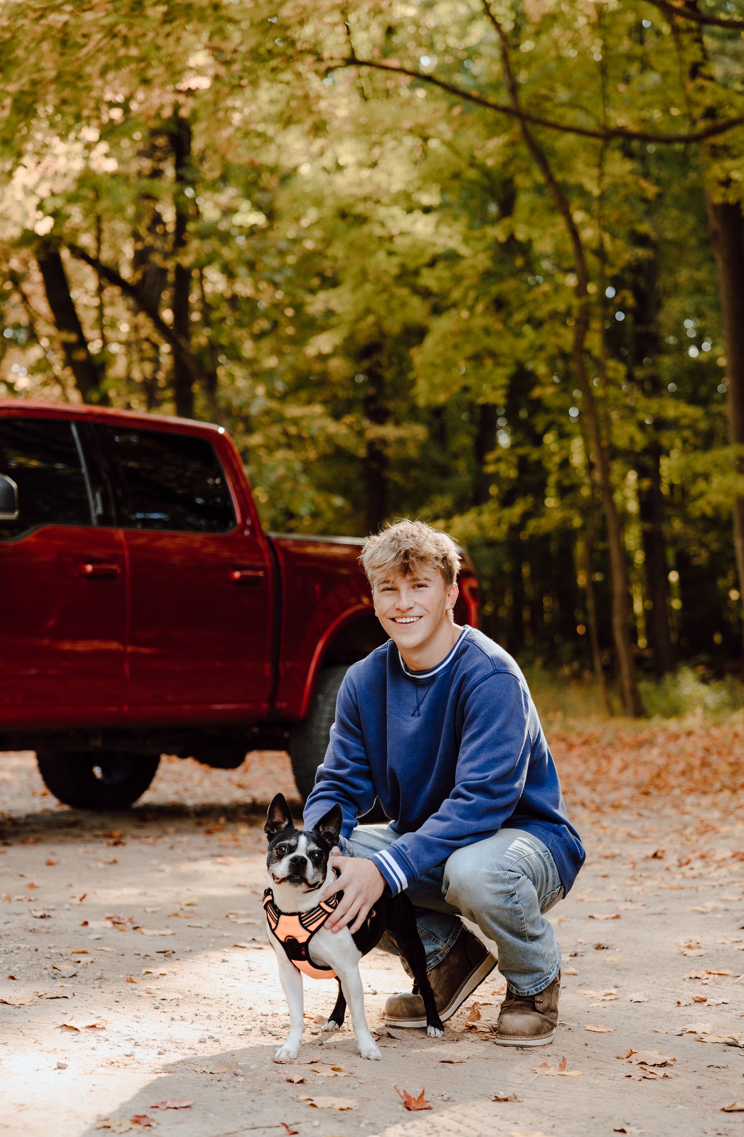 Person in blue sweater sitting beside a red truck with a small black and white dog.