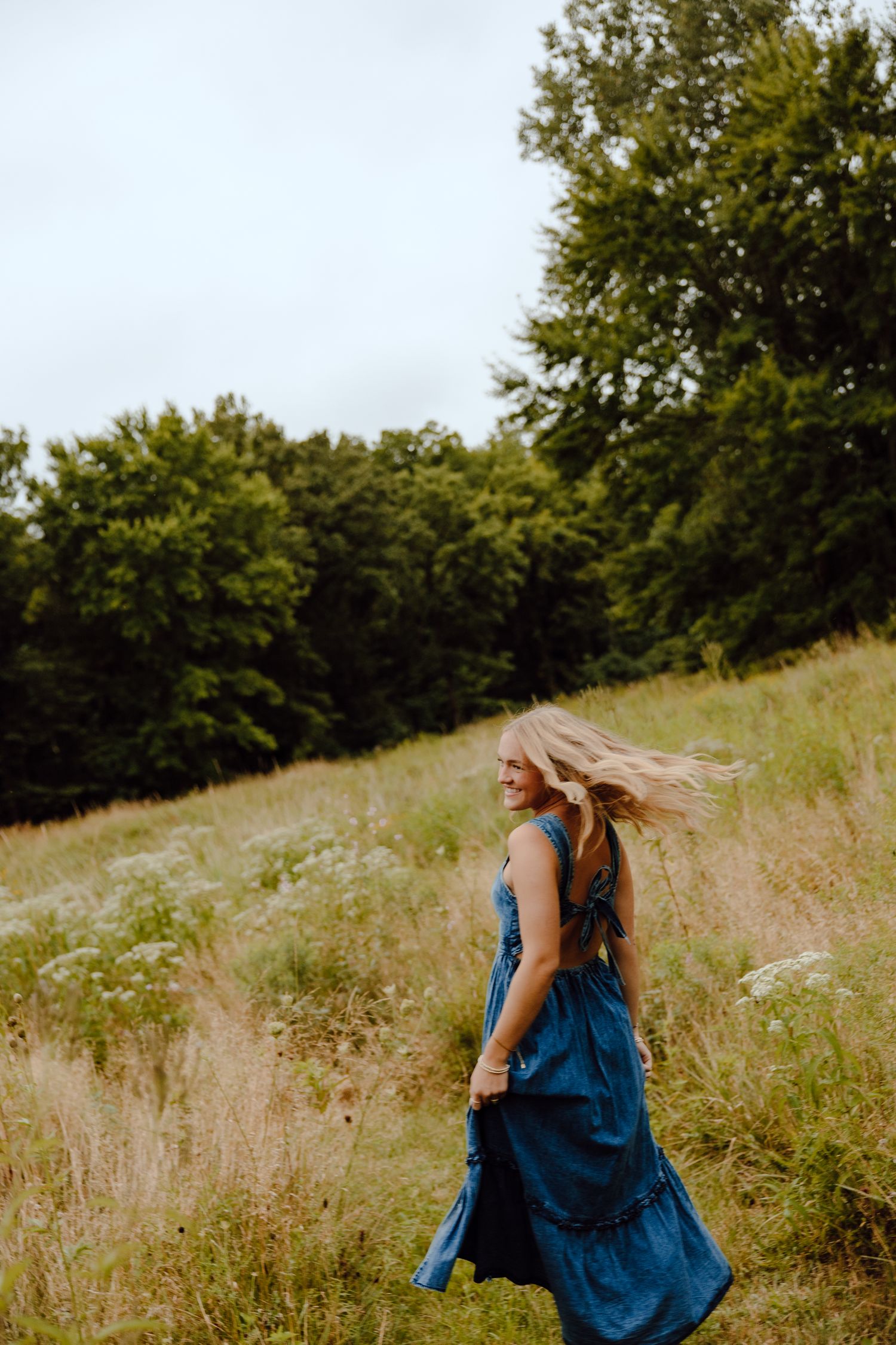 Woman in blue dress twirls joyfully in a sunlit meadow with trees in the background.