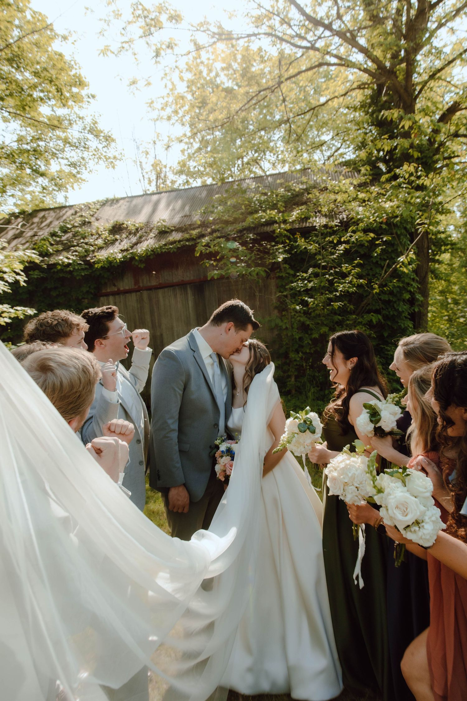 Bride and groom share first kiss surrounded by cheering wedding party outdoors near rustic barn in sunlight.