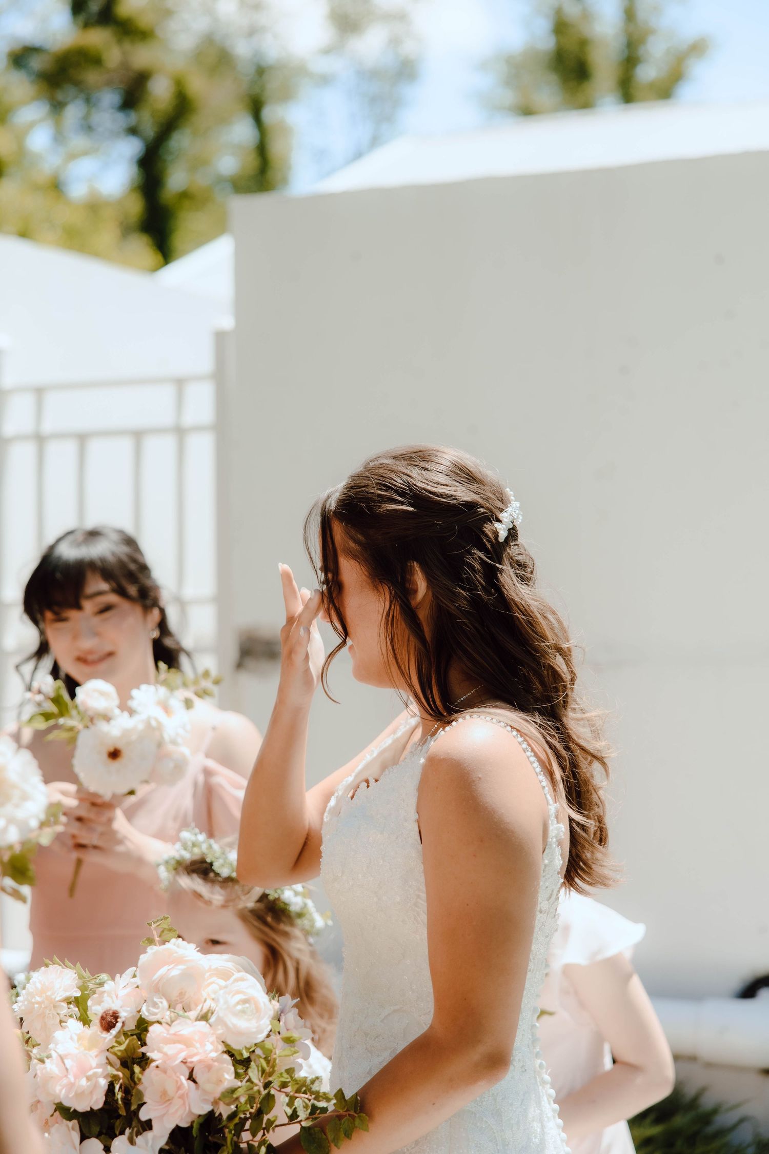Emotional bride in white lace gown wiping tears while bridesmaid in pink holds bouquet of white roses outdoors.