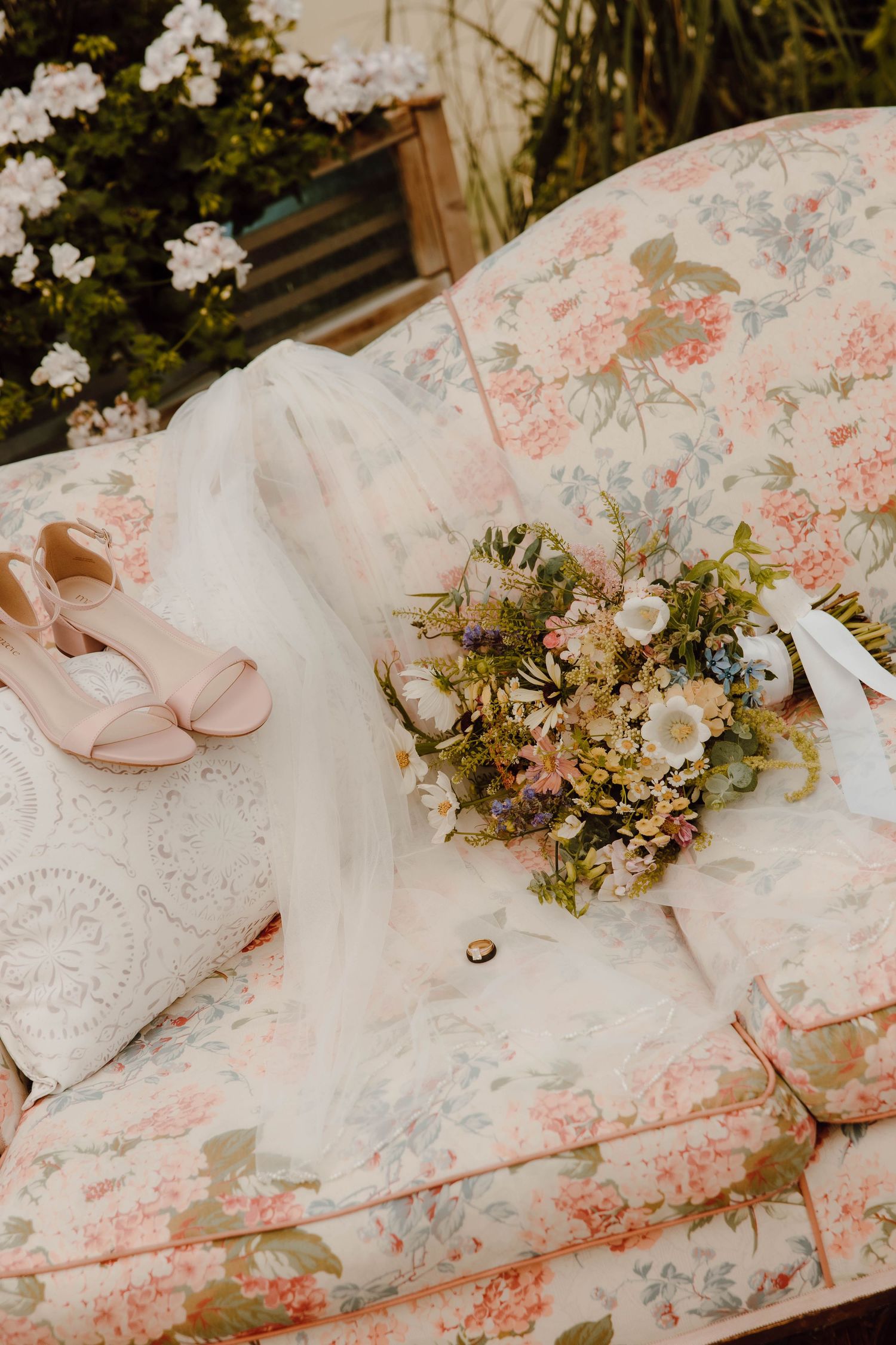 Bridal details on floral sofa with blush heels, wildflower bouquet, white veil, and wedding ring.