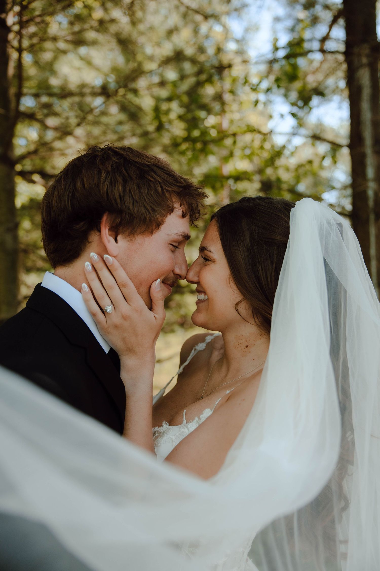 A joyful bride and groom share a romantic nose-to-nose moment outdoors, her veil flowing as she touches his face.