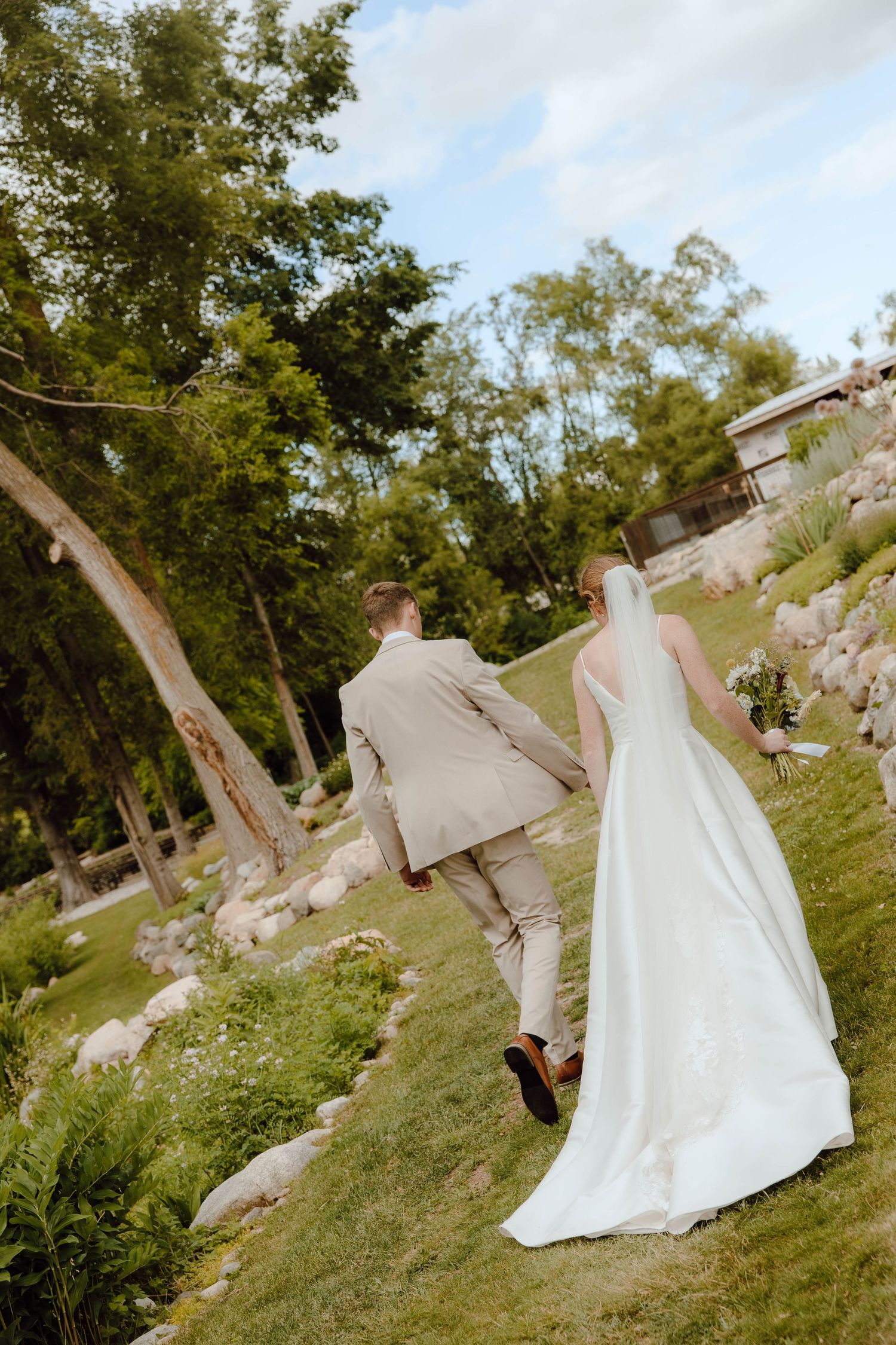 Bride and groom walking hand in hand through a scenic garden, bride holding a bouquet in a white gown.