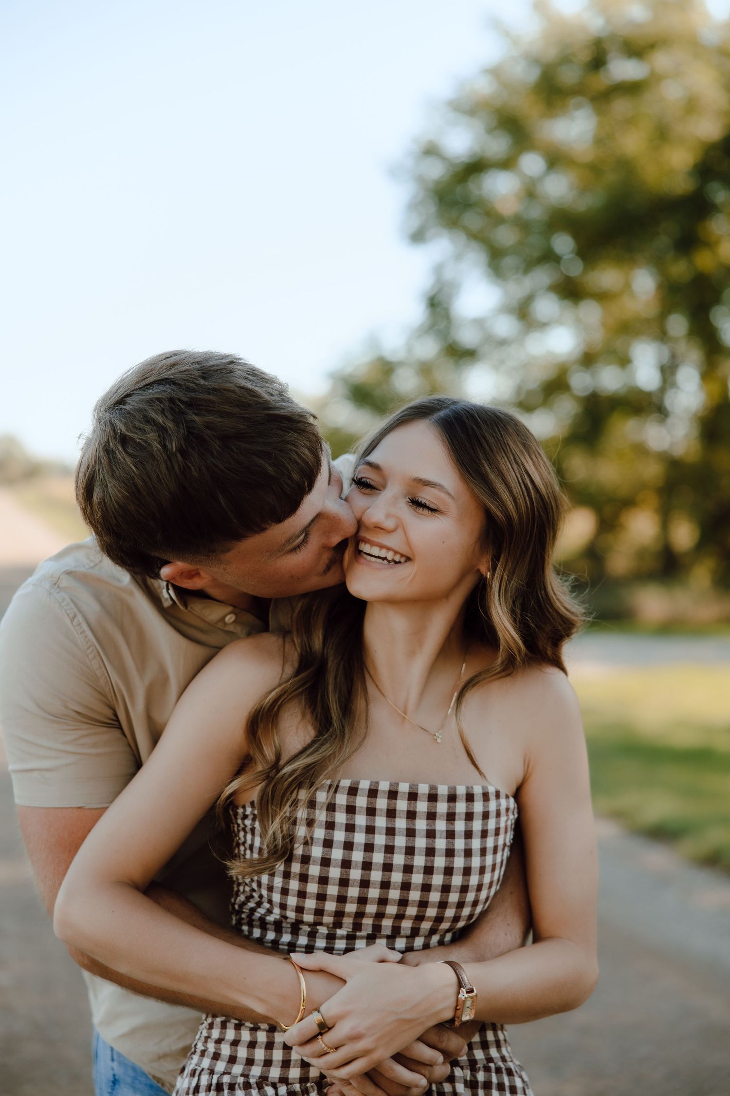 A happy couple shares a romantic kiss outdoors, with the woman smiling joyfully in a checkered dress.