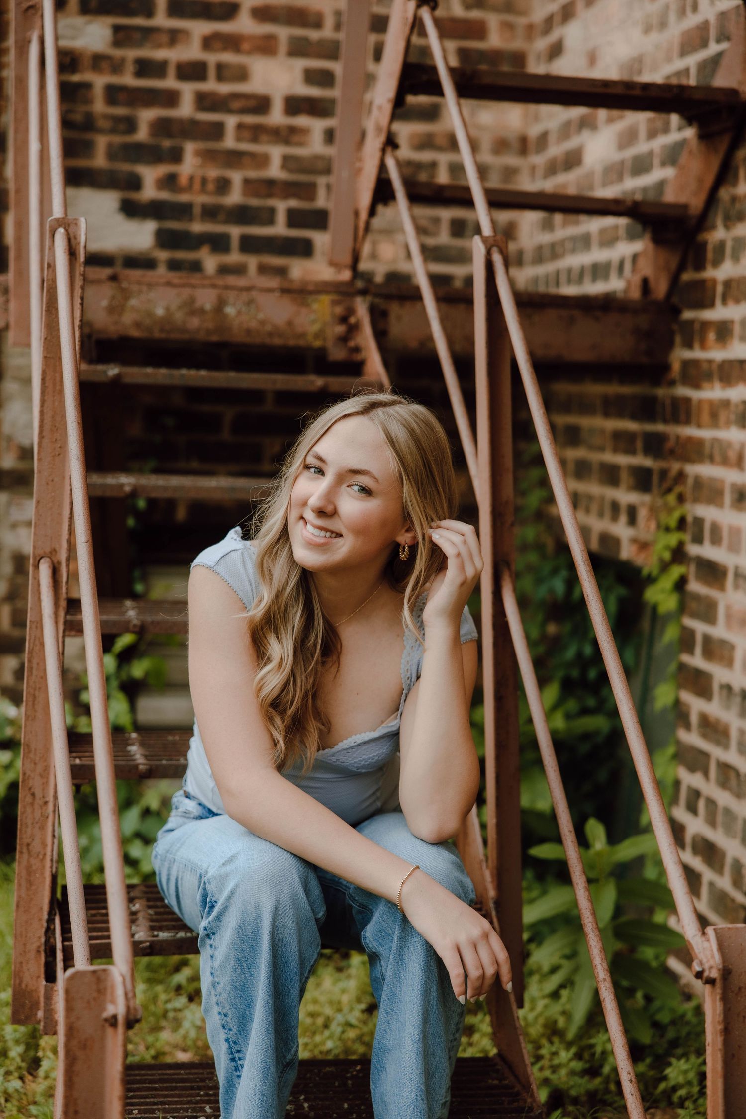 Young woman in blue jeans and top sitting on rustic iron stairs against a brick wall, smiling softly.