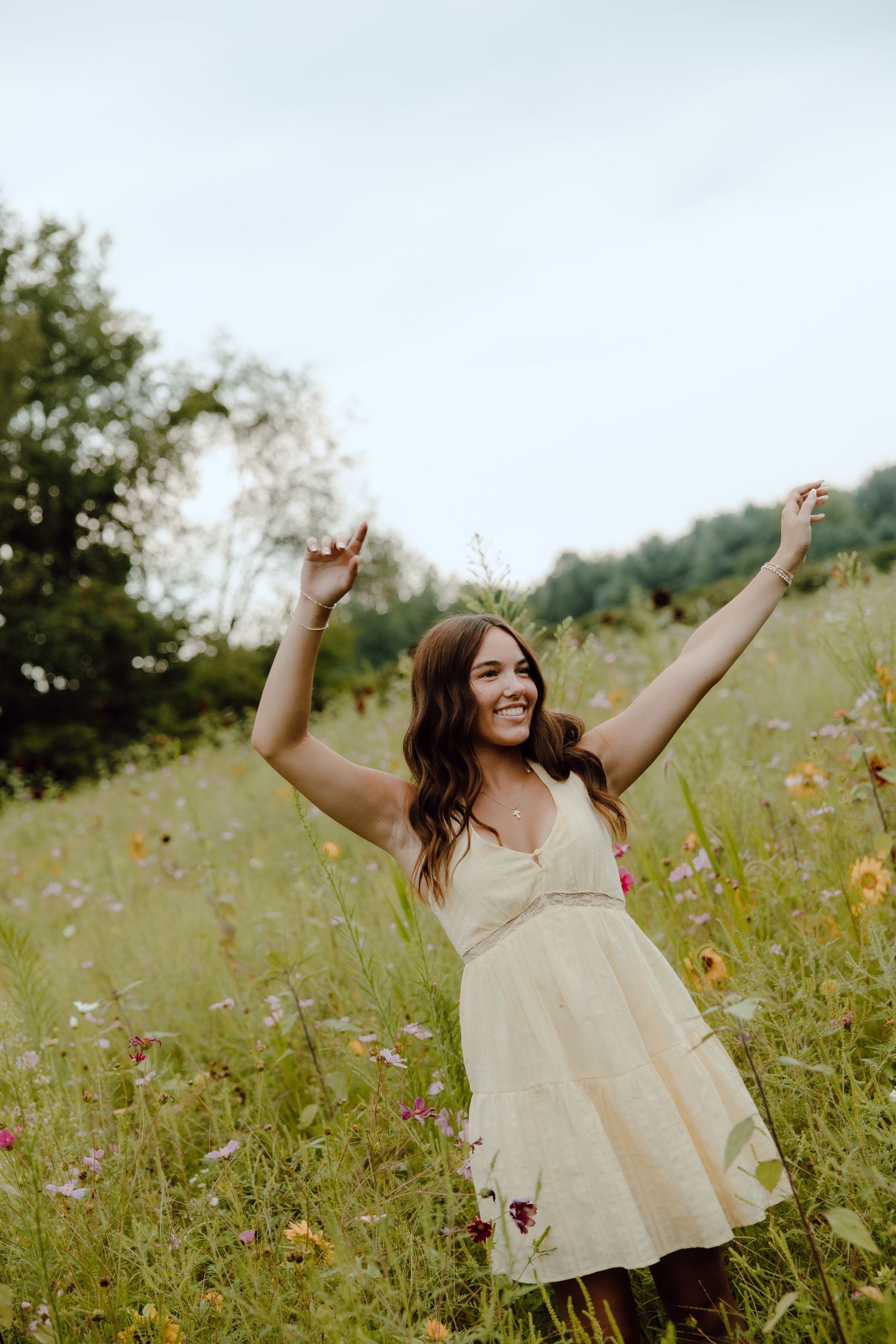 Joyful young woman in floral dress with arms raised in a vibrant wildflower field on a cloudy day.