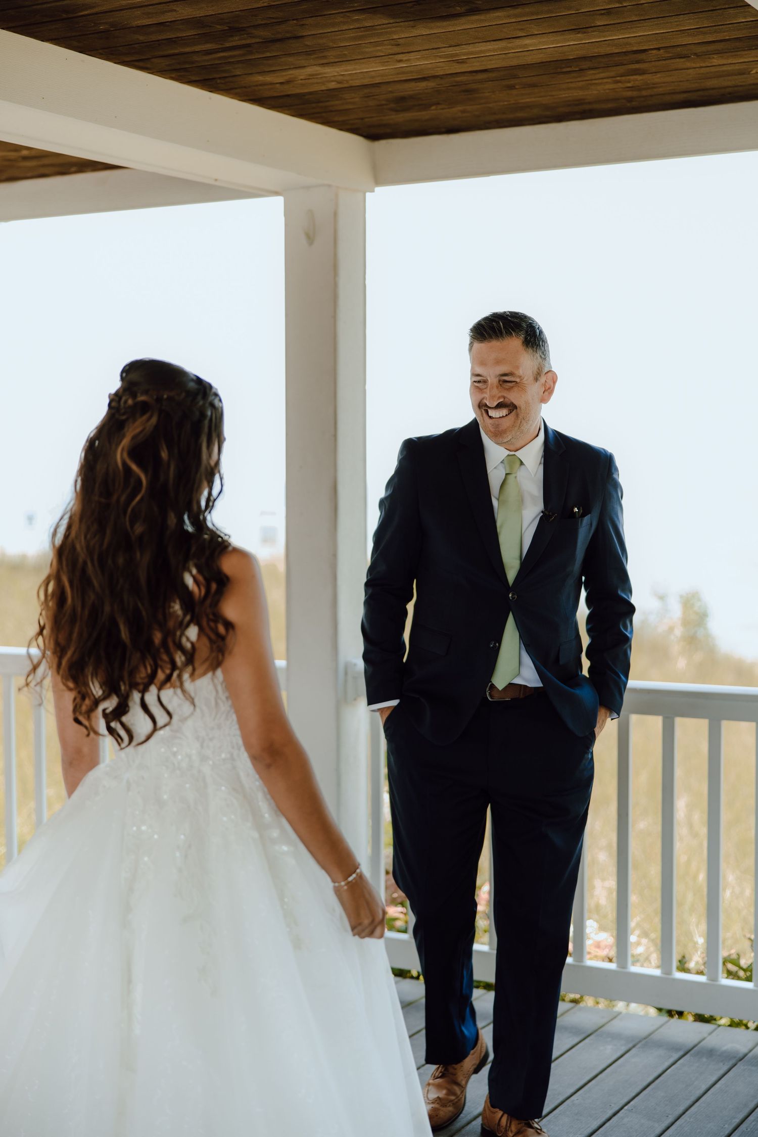 Groom in navy suit smiles at bride in white wedding gown on a sunlit porch during first look.