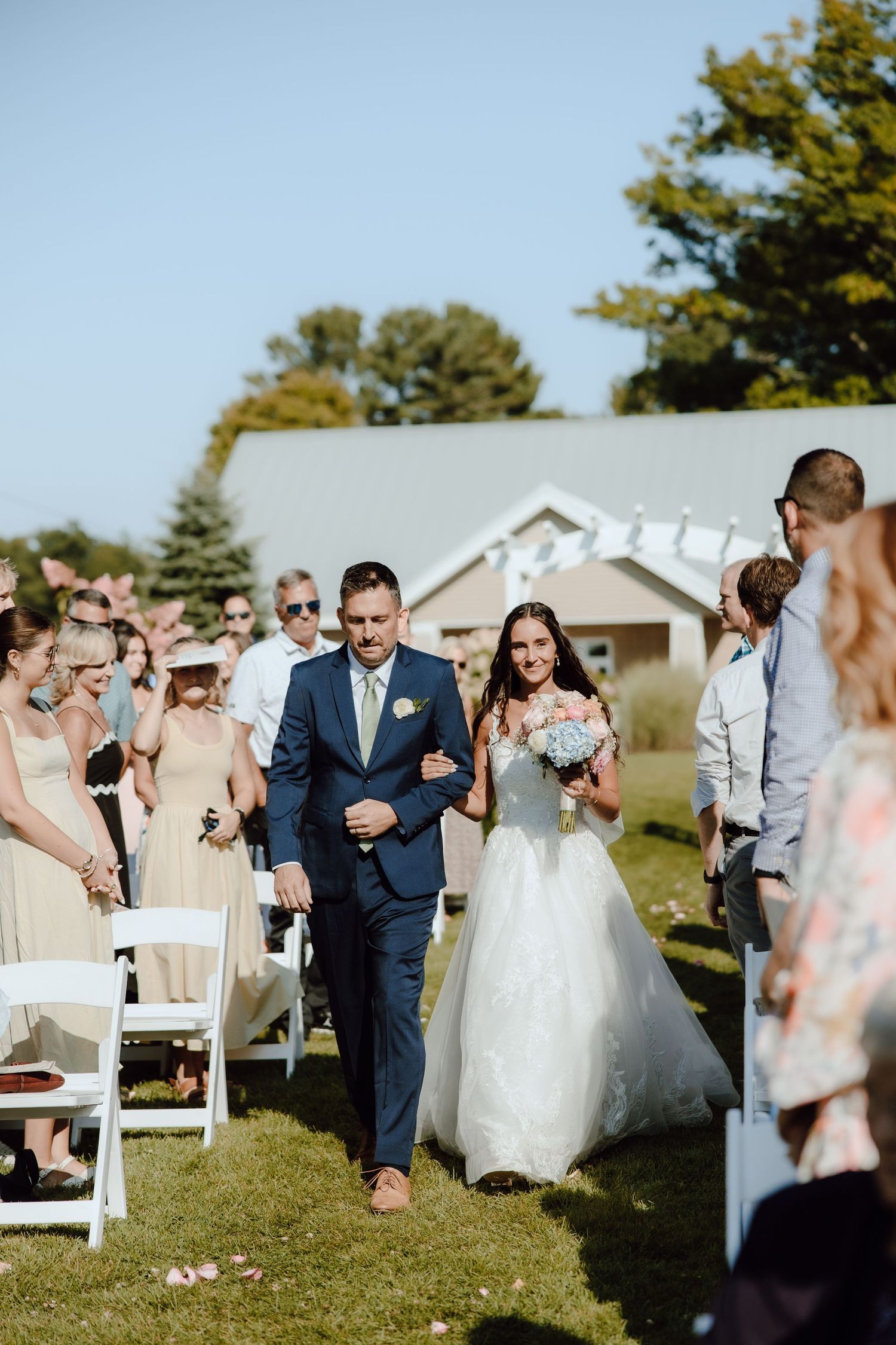 Bride walking down outdoor aisle with escort holding pink bouquet at sunny garden wedding ceremony.