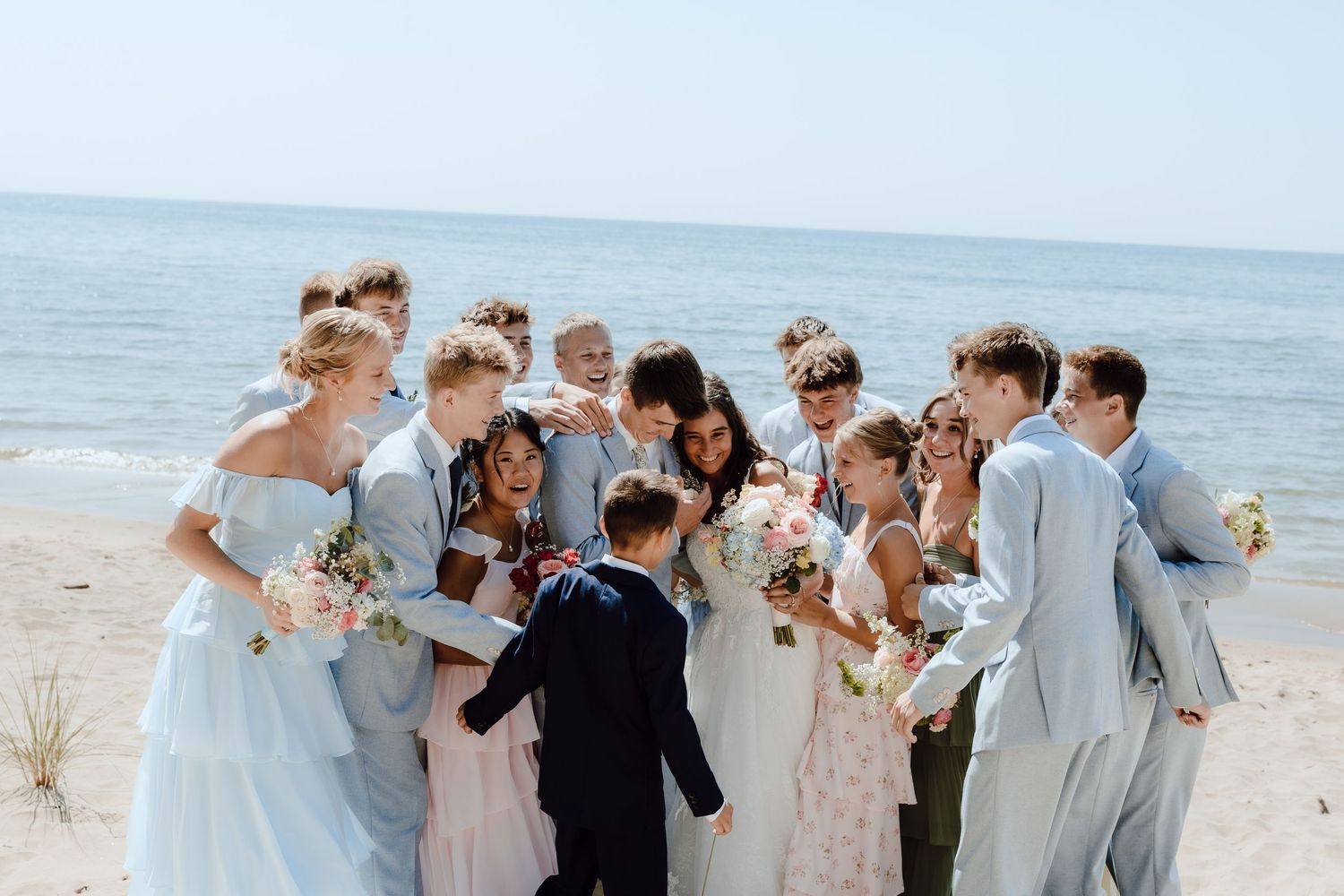 Wedding party group hugging and celebrating together on a sunny beach with blue ocean in background.