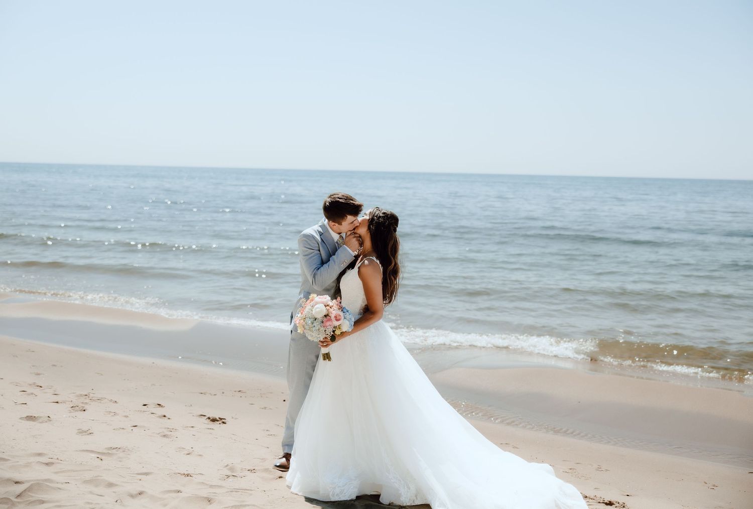 Bride and groom share a romantic kiss on a sandy beach with calm ocean waves in the background.