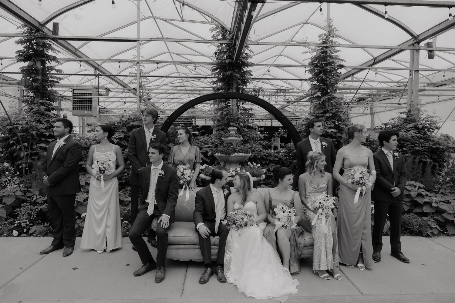 Black and white wedding party photo in a greenhouse with bride, groom, and attendants near a fountain.