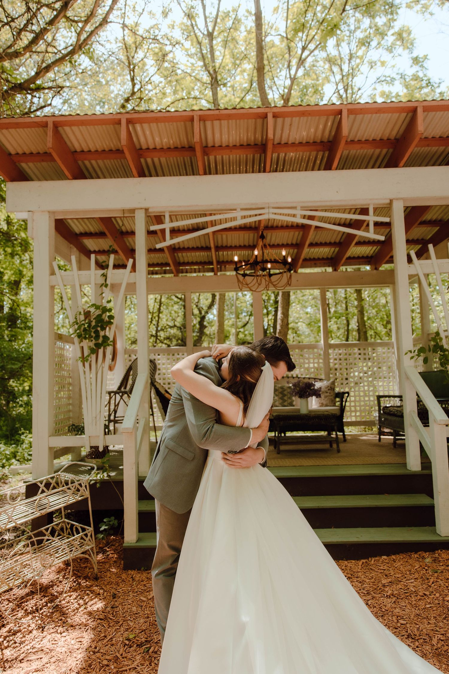 A bride and groom share a romantic embrace outside a charming rustic gazebo surrounded by lush green trees.