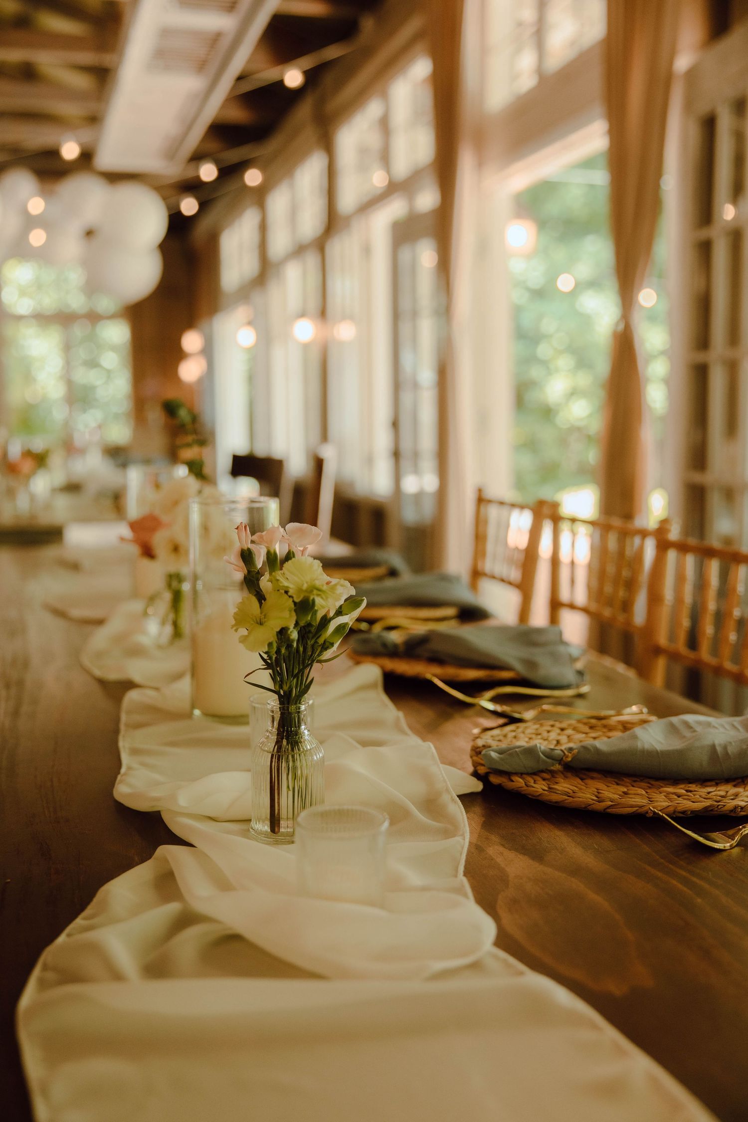 Elegant wedding reception long table with white runner, small floral centerpieces, wicker chargers, and string lights in a rustic venue.