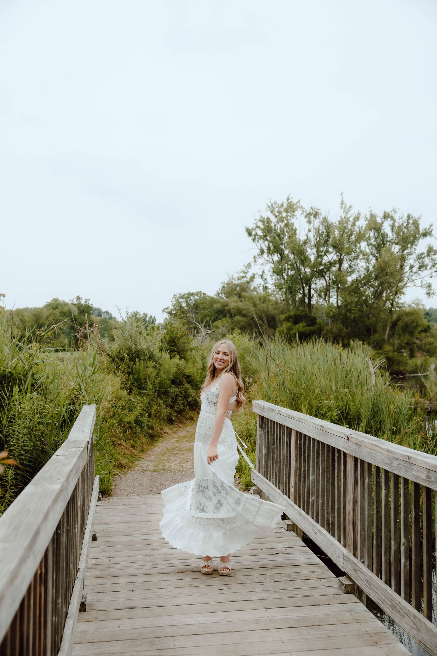 Blonde woman in white lace dress twirling on wooden boardwalk surrounded by lush green vegetation.