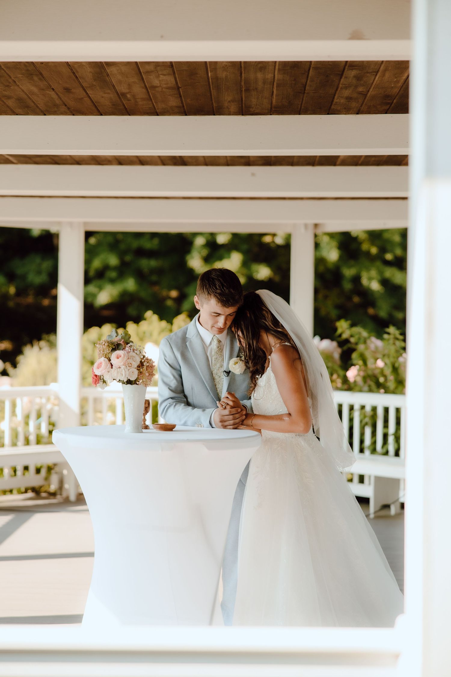 Bride and groom share a tender moment at a white cocktail table in an elegant outdoor gazebo setting.