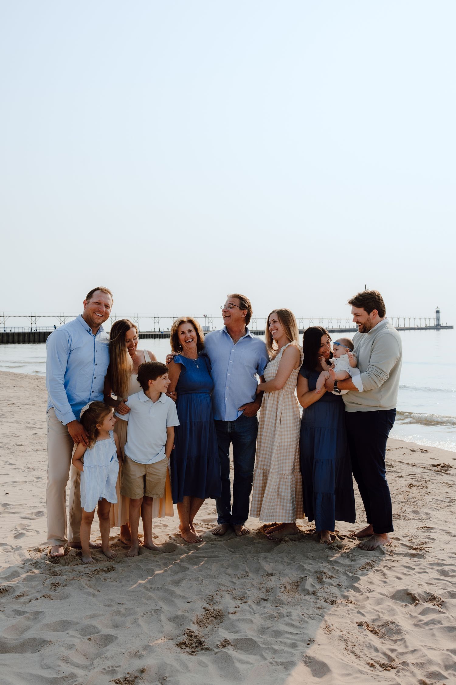Extended family gathering for a group photo on a sandy beach with a pier in the background.