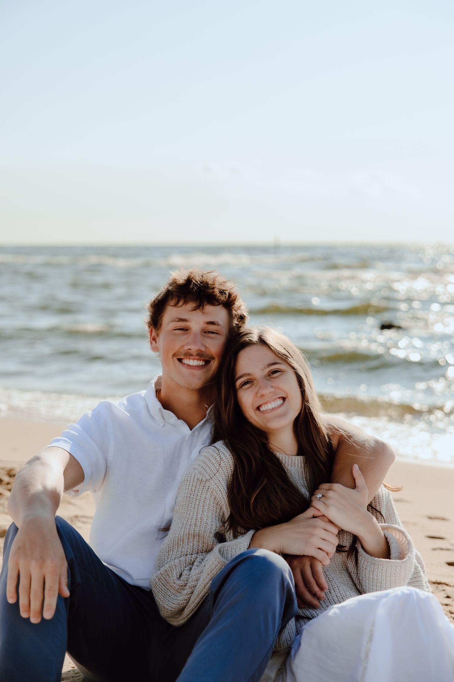 A couple sits together on a beach smiling as waves crash behind them on a sunny day.