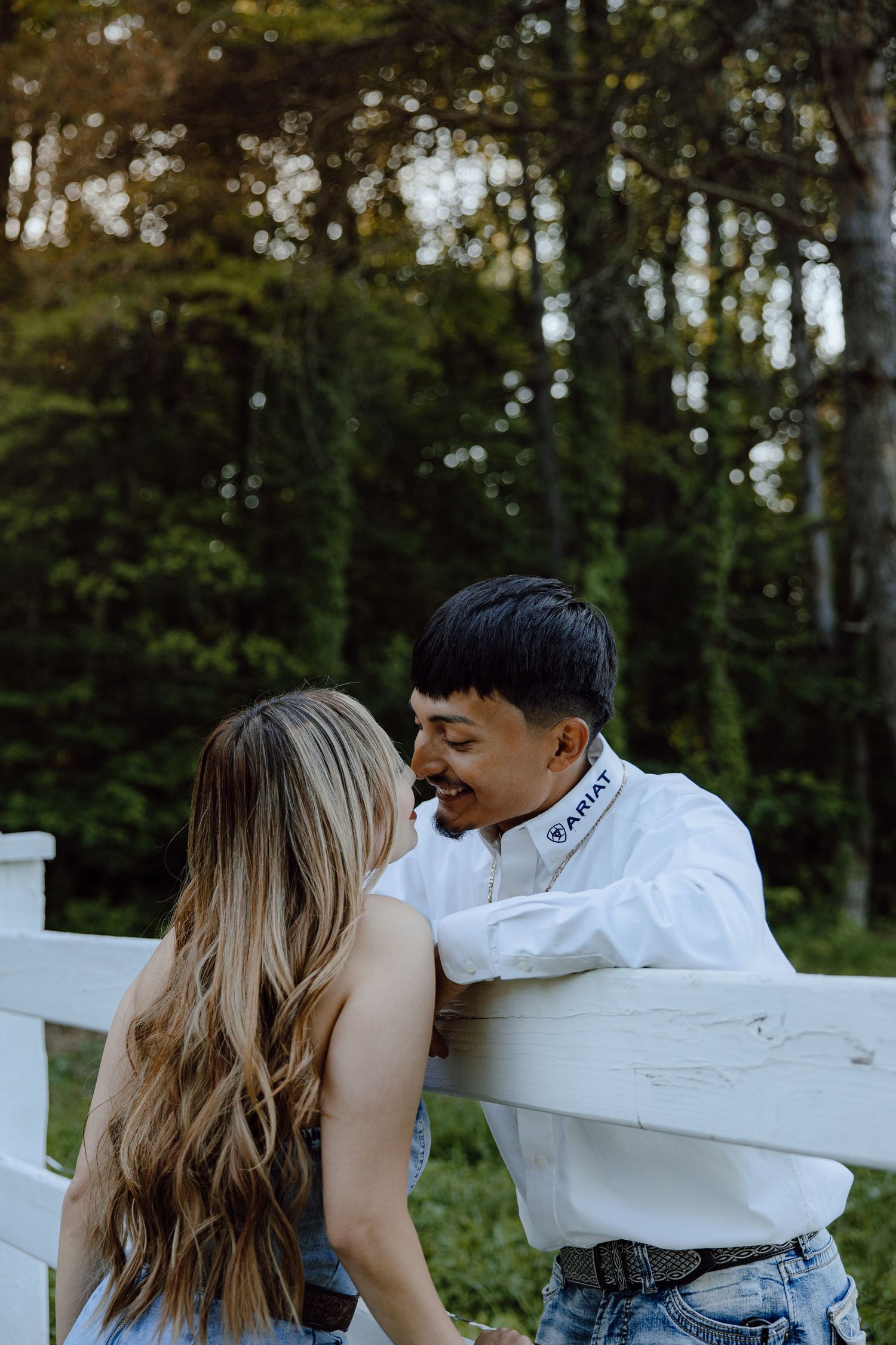 Romantic moment shared between couple leaning on white fence in wooded setting.