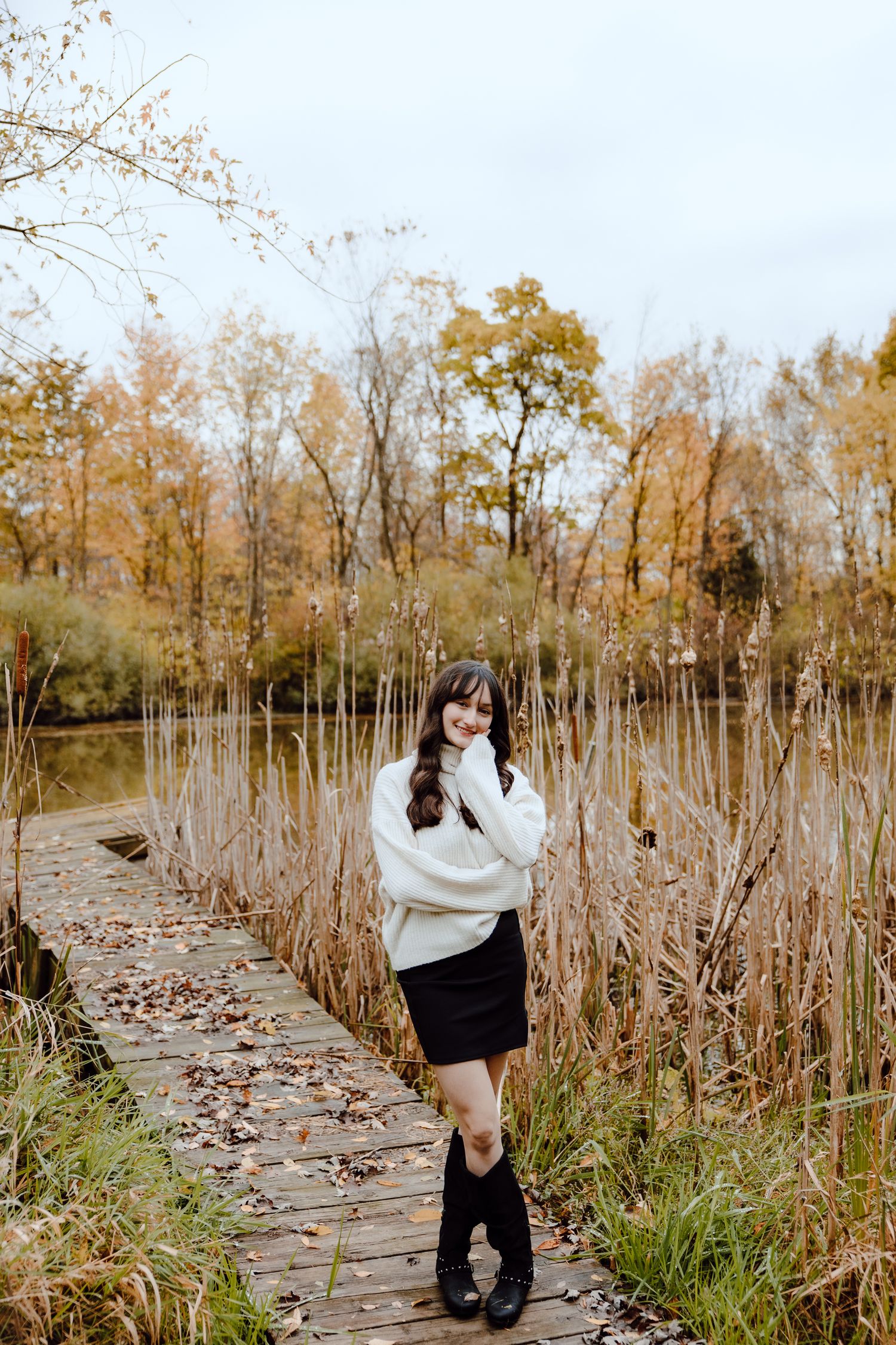 Person in white sweater and black skirt poses playfully on wooden boardwalk surrounded by autumn reeds.