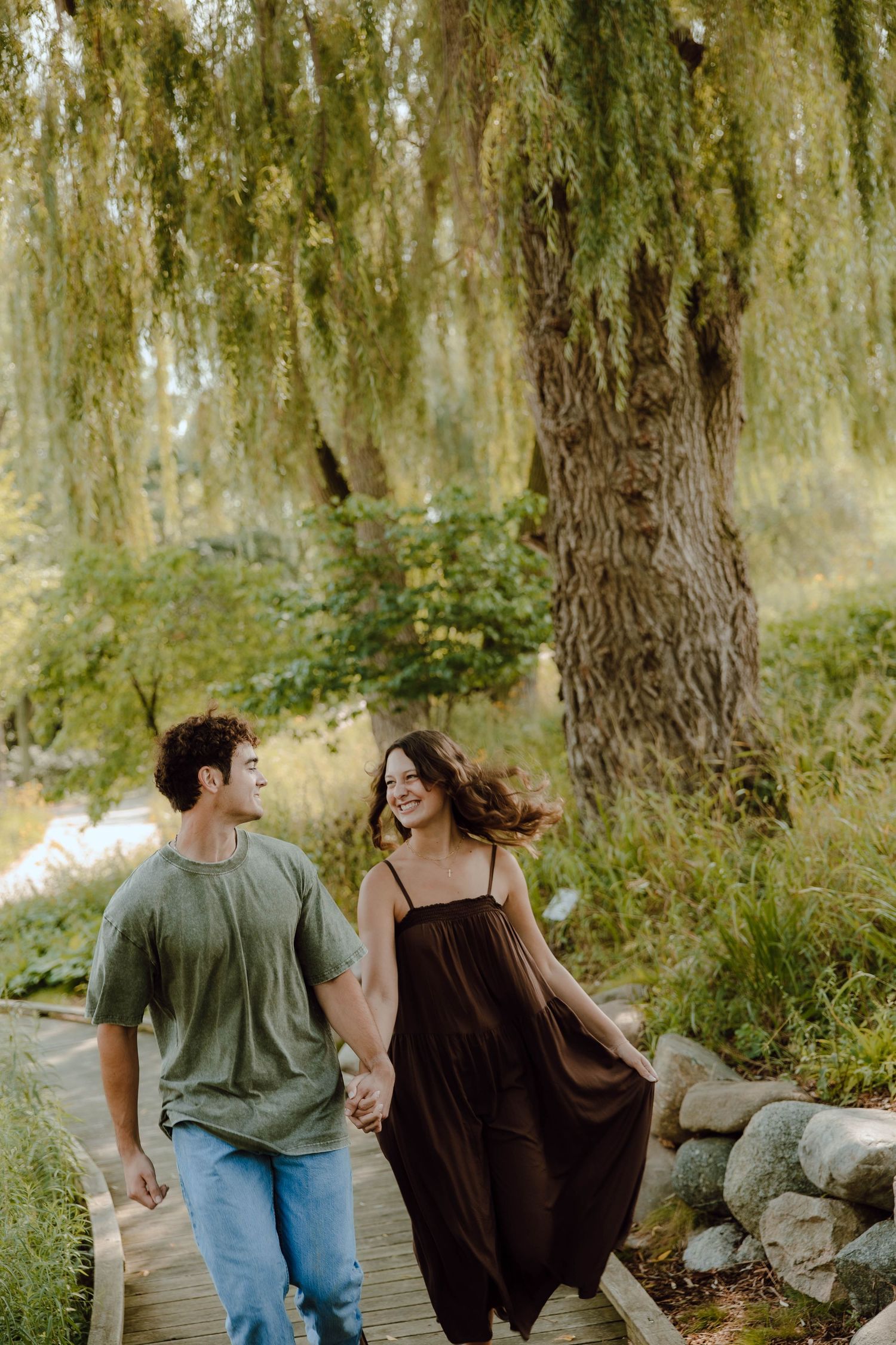 A couple walks hand-in-hand on a wooden path under a weeping willow tree, smiling at each other.