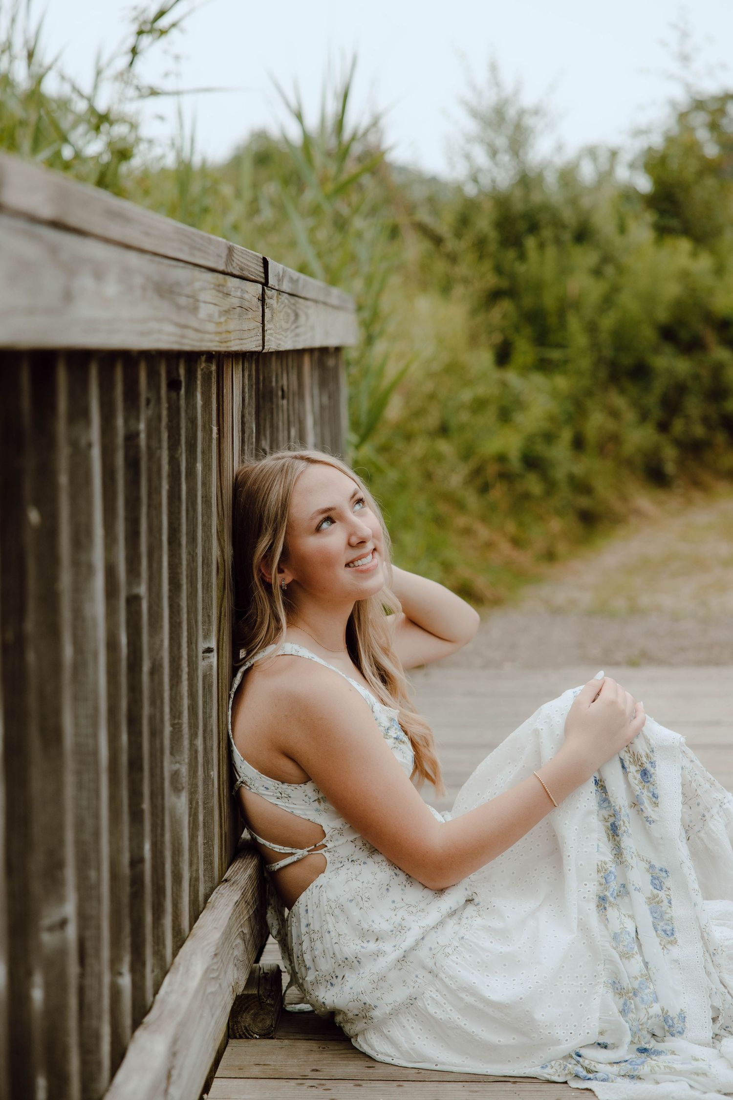 Blonde woman in white floral dress sitting on wooden boardwalk leaning against fence in nature.