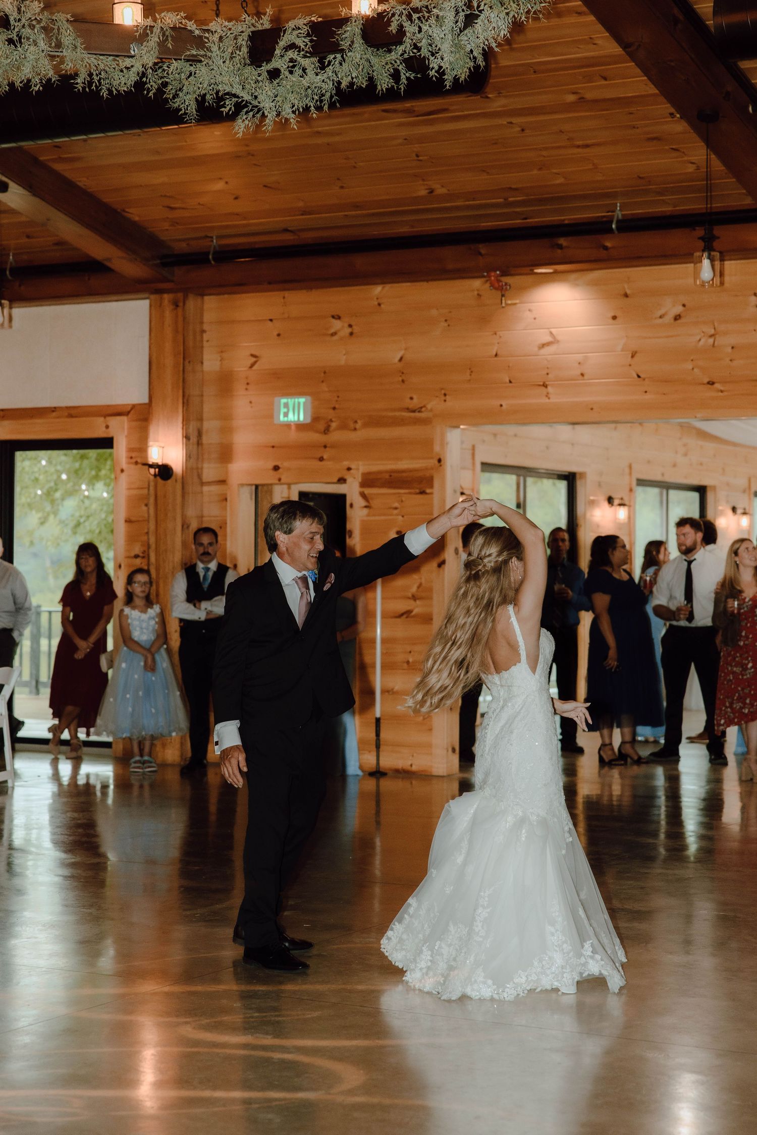Bride in lace gown dancing with man in black suit at rustic wood wedding venue.