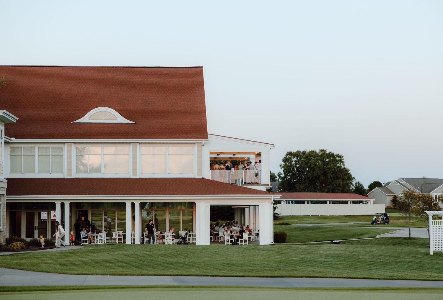 Wedding reception venue at dusk with guests gathering on patio of large clubhouse building.