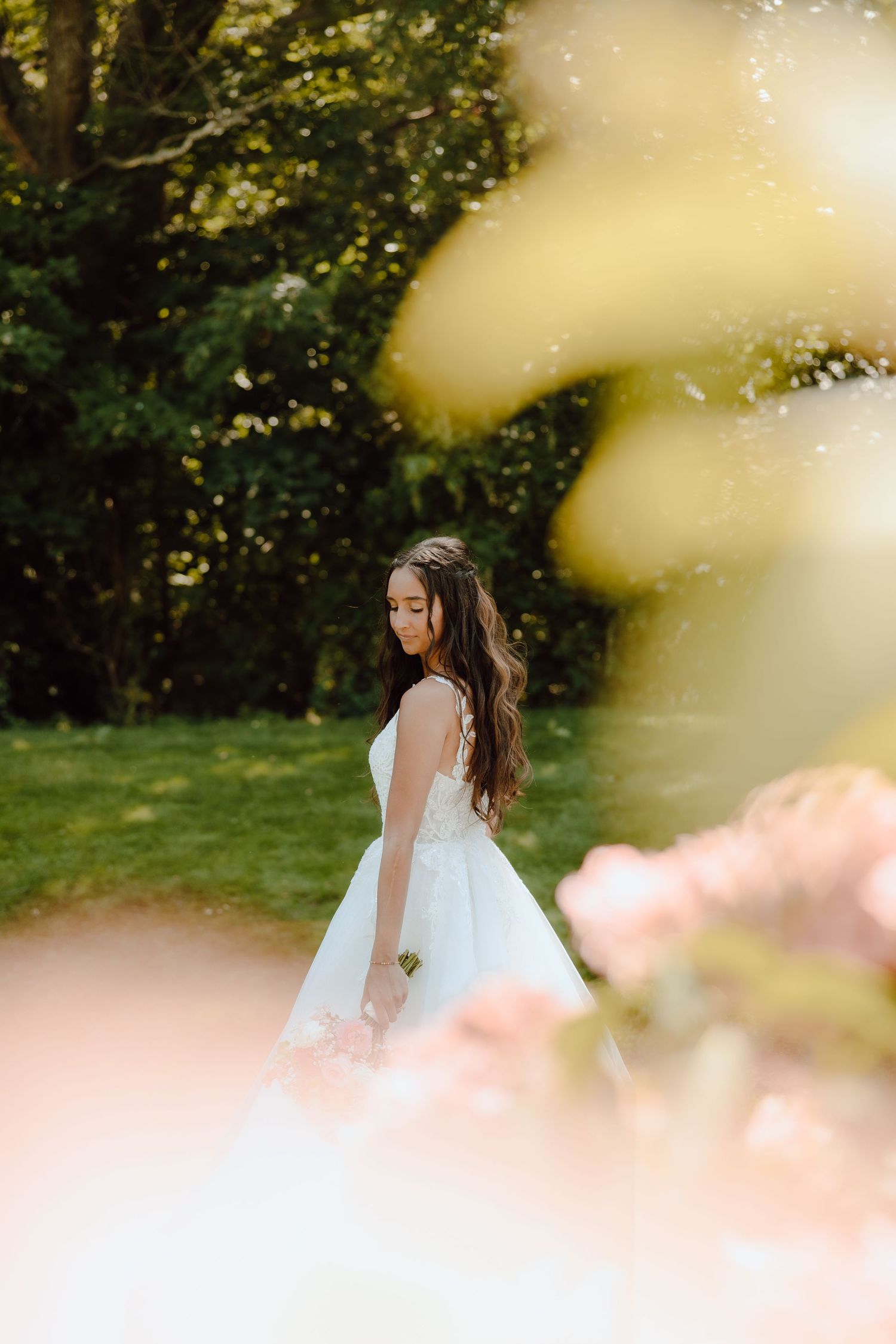 A bride in a white ball gown stands gracefully outdoors, surrounded by lush greenery and soft bokeh flowers.
