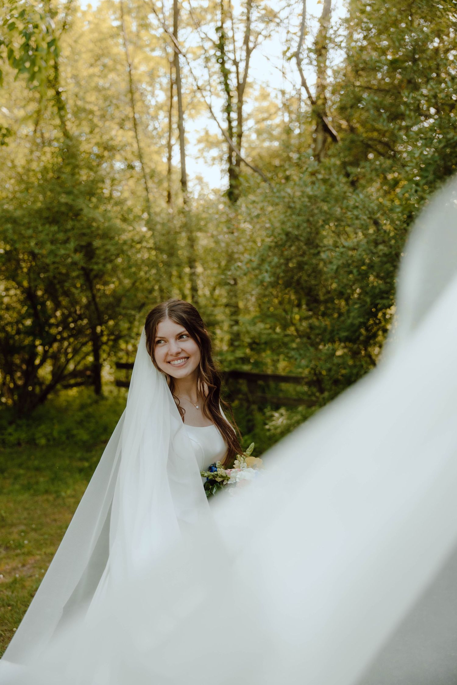 Radiant bride in a flowing white gown and veil smiles beautifully amid lush green trees at an outdoor wedding.