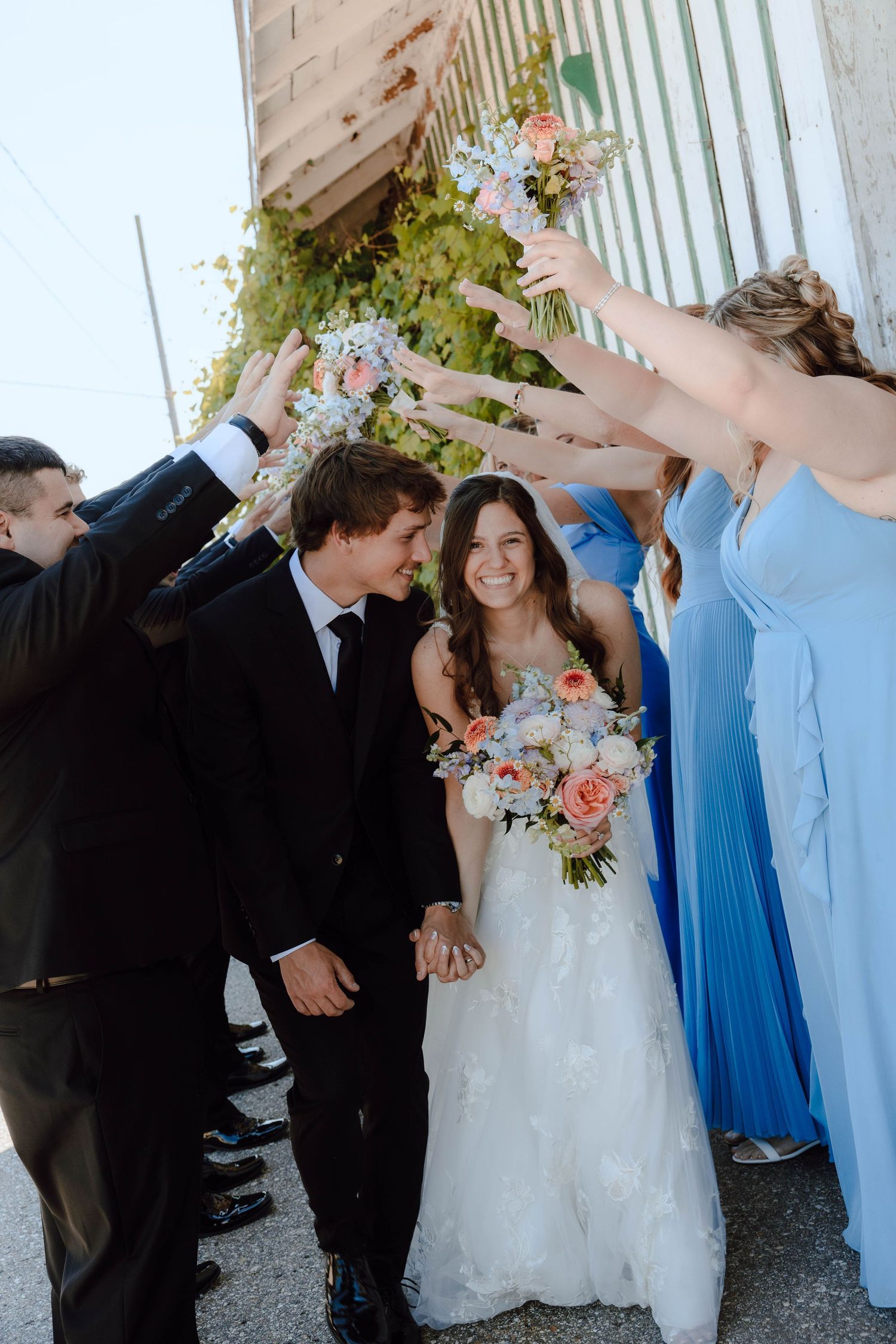 Bride and groom walk through arch of raised bouquets, bridesmaids in blue dresses, outdoor wedding celebration.