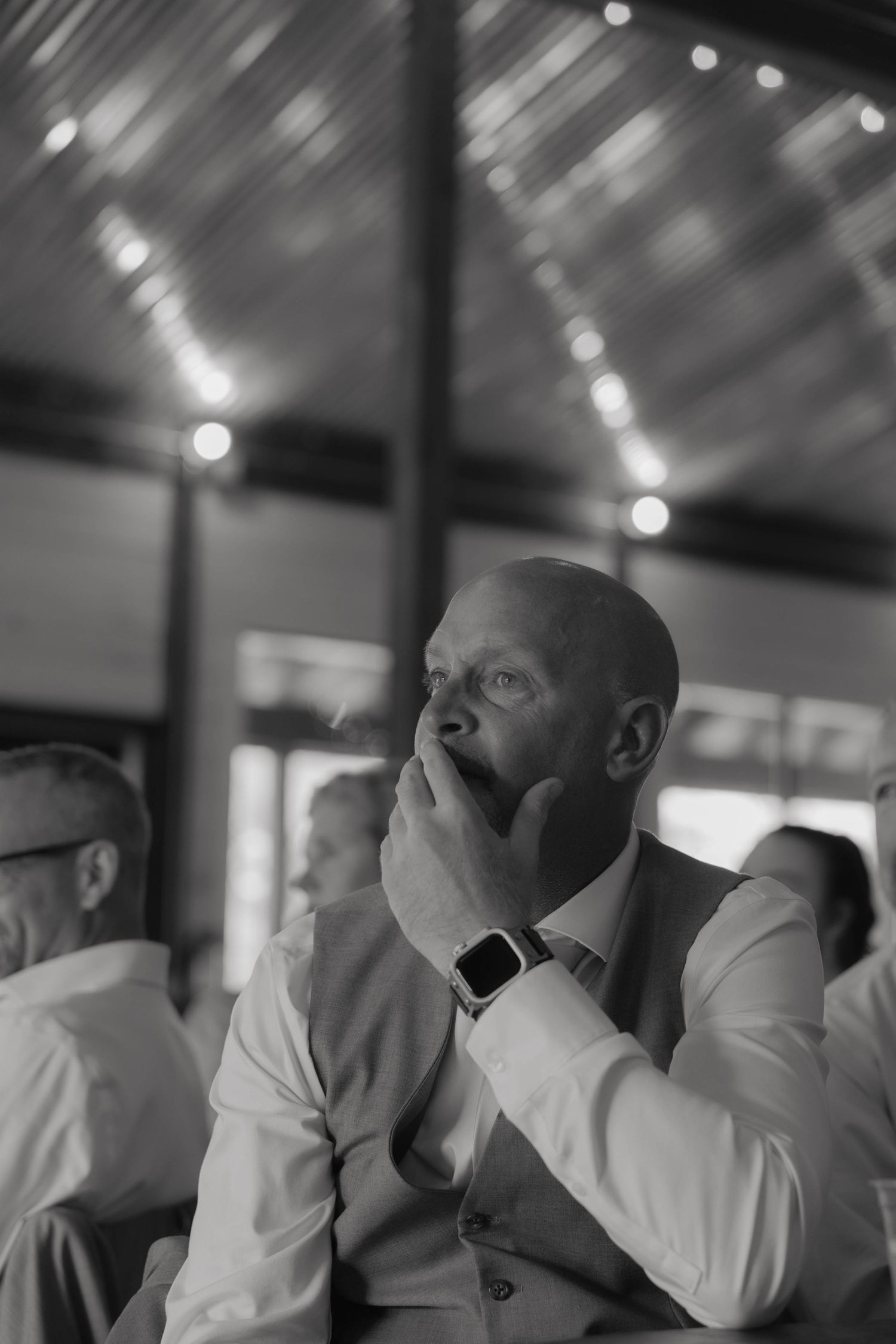 Black and white photo of emotional bald groom covering mouth with hand during wedding ceremony, wearing vest and smartwatch.