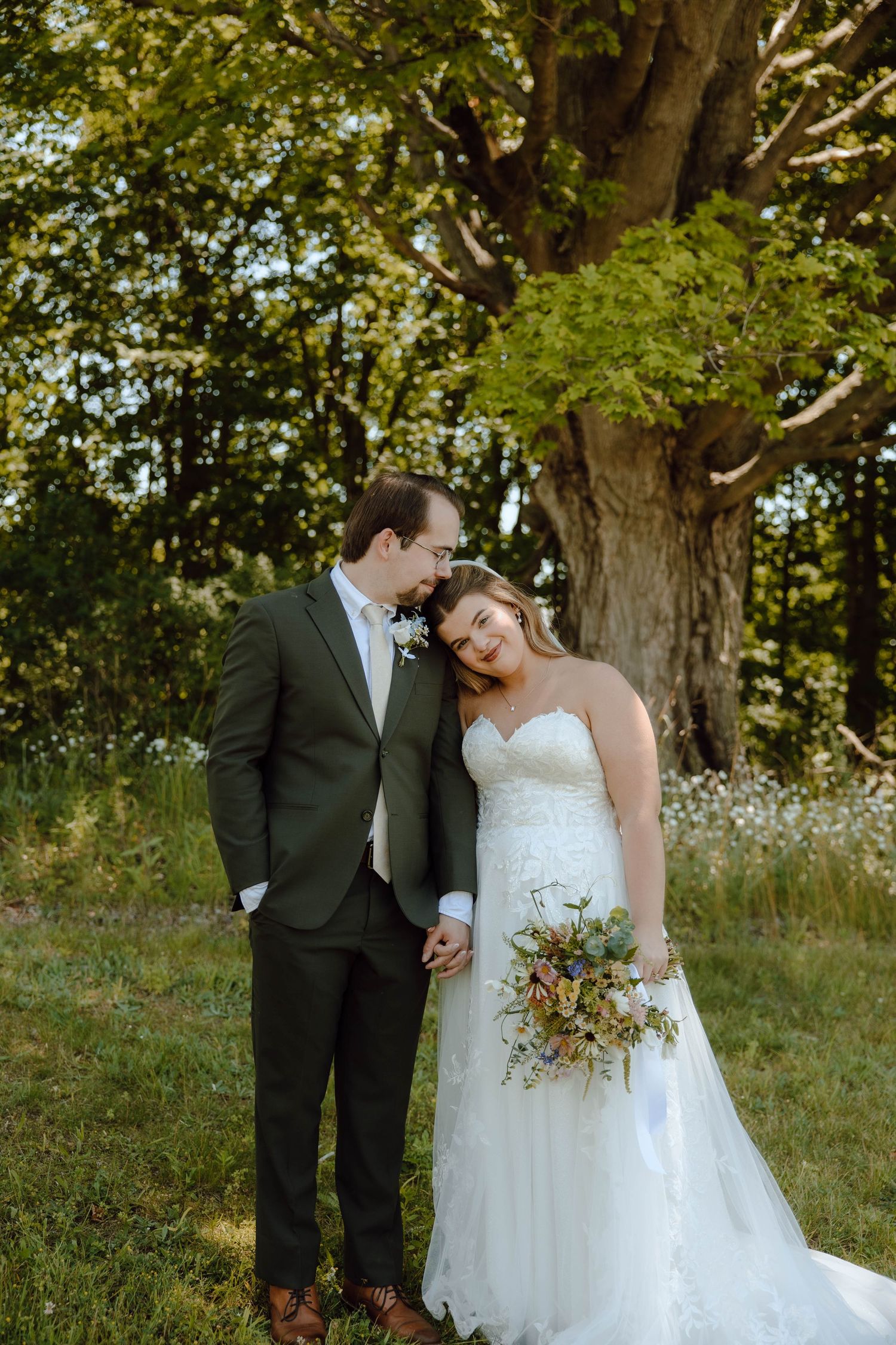 Bride and groom share a tender moment under a large oak tree, bride holding a wildflower bouquet in a lace gown.
