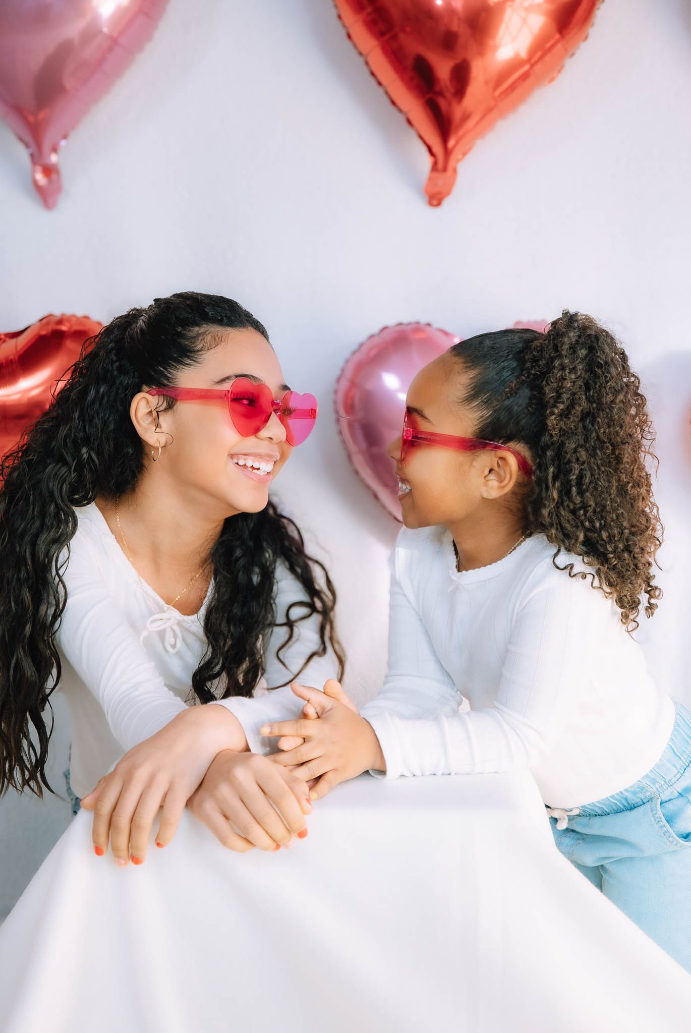 A woman and young girl wearing heart-shaped sunglasses smile at each other near pink and red heart balloons.