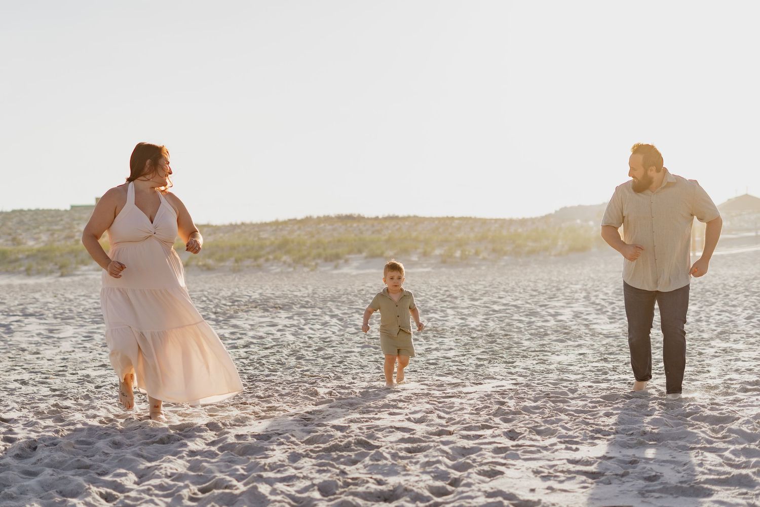 Family of three enjoying a sunny beach day, with a toddler running between parents on white sand.