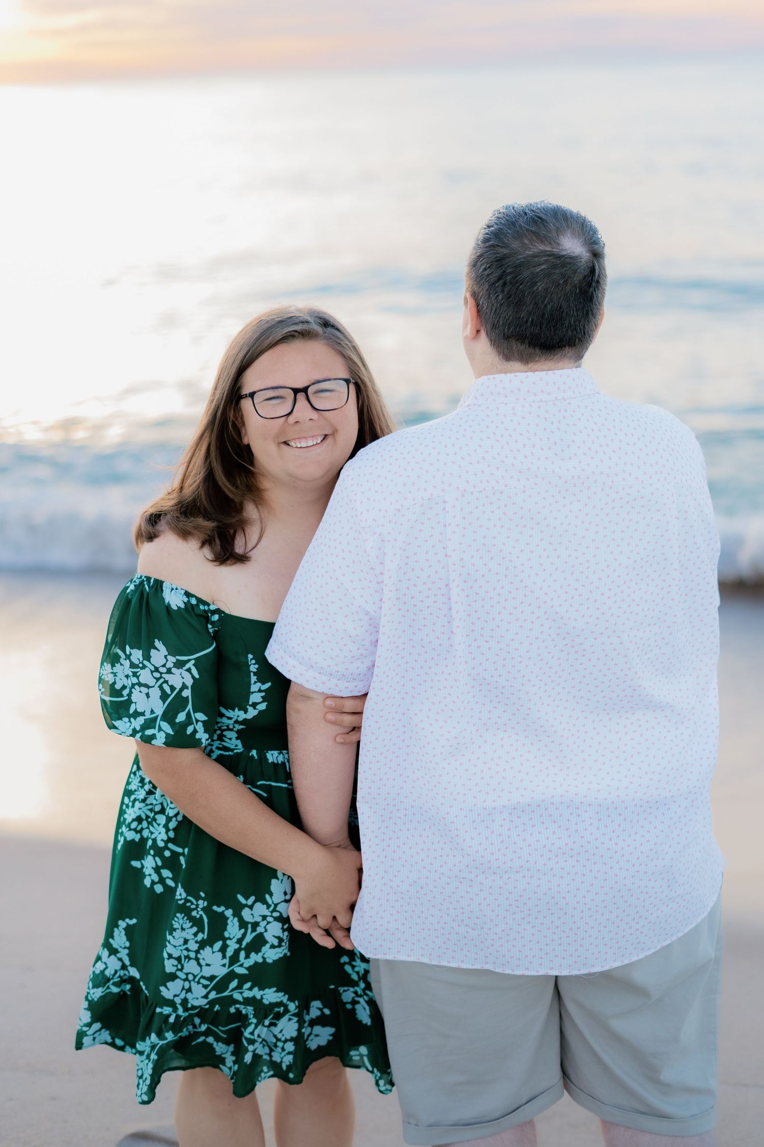 Couple holding hands on beach at sunset, woman in green floral off-shoulder dress smiling at camera, man in white shirt.