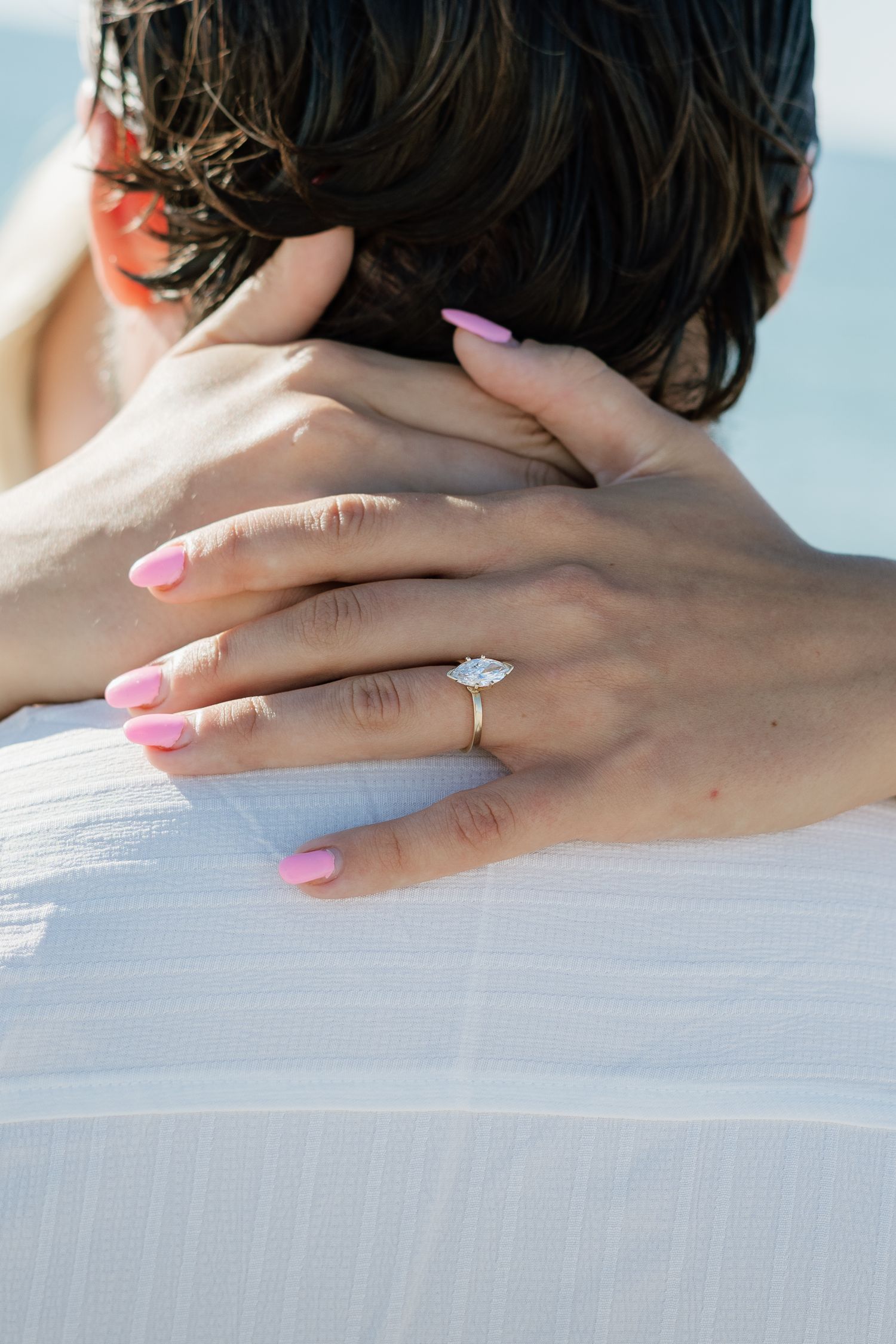 Close-up of a woman's hand with pink nails and a diamond engagement ring resting on a man's back at the beach.