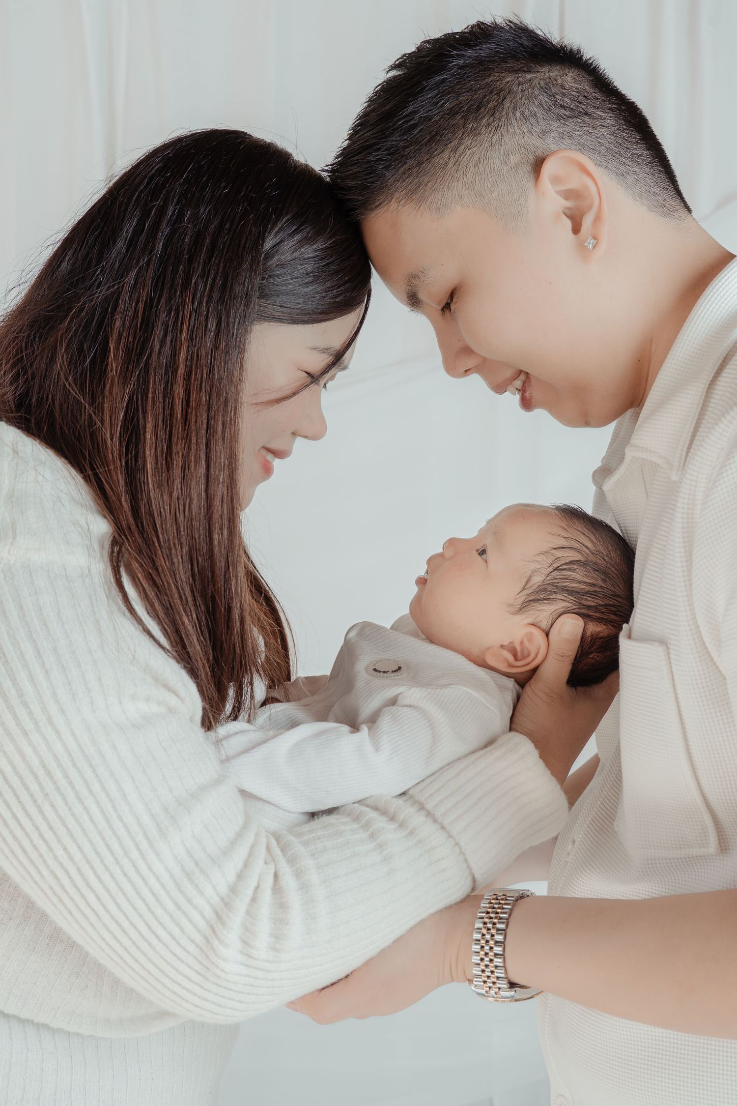 Asian parents tenderly holding their newborn baby, smiling lovingly in a soft white studio setting.