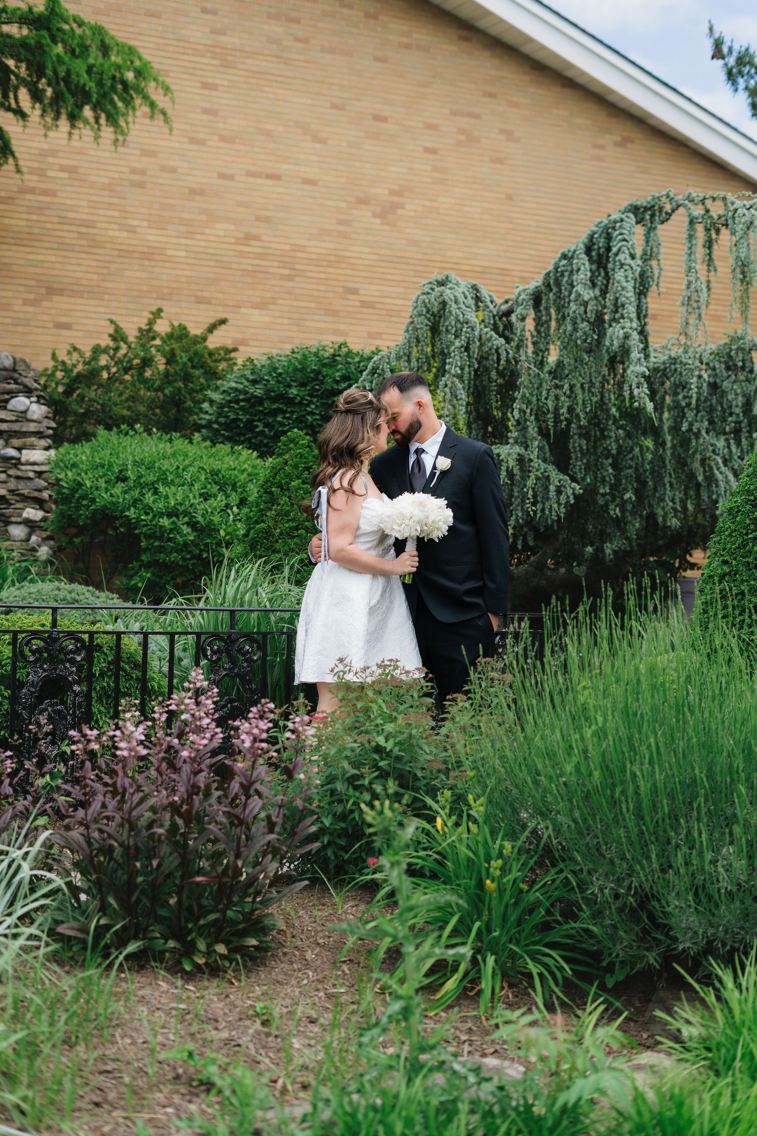 Newlywed couple shares a tender kiss surrounded by lush garden greenery outside a brick church.