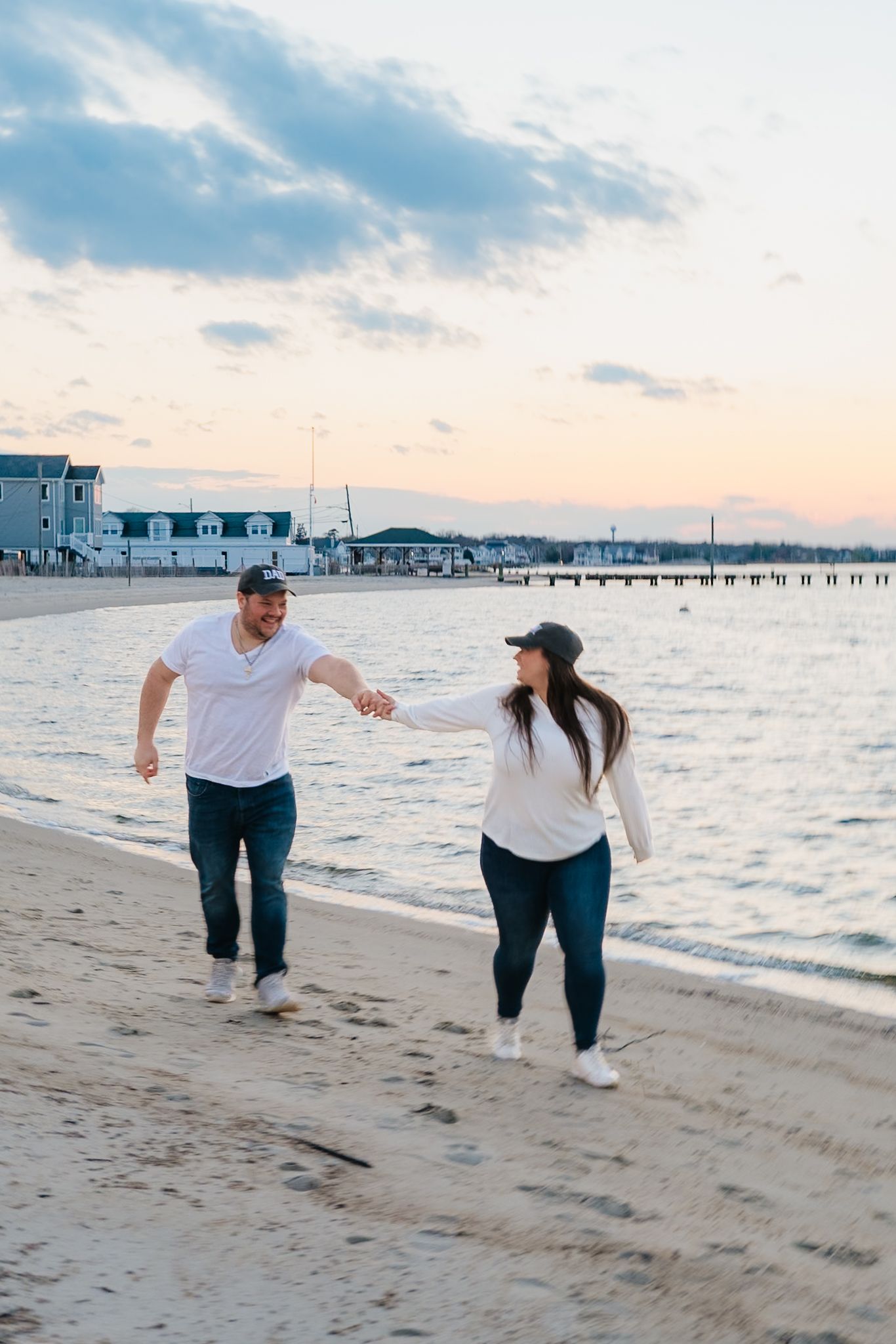 Couple holding hands and walking along a sandy beach at sunset with a pier and waterfront homes in the background.