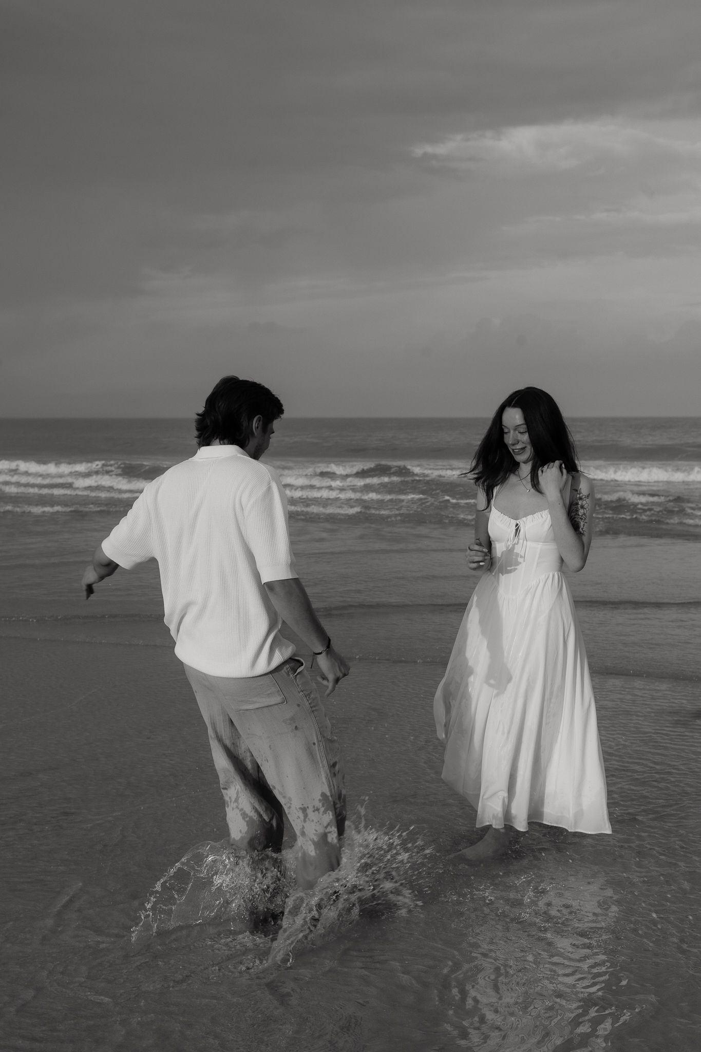 Black and white photo of couple playing in ocean waves, woman in white dress laughing on beach.